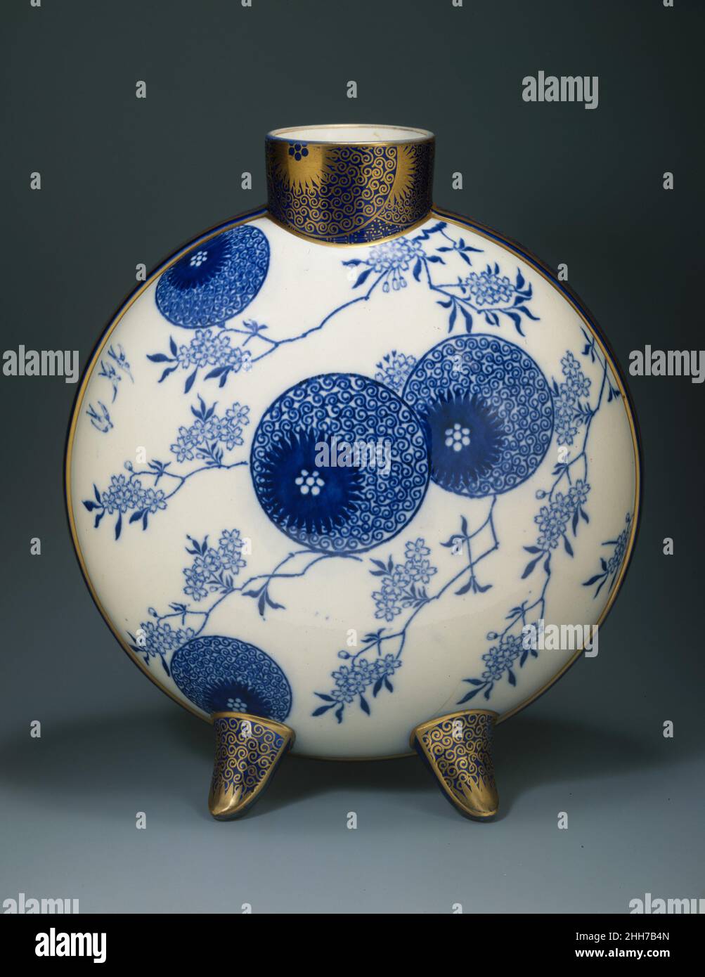 Mondflasche ca. 1878–82 Designed by Henry Slater Diese Vase ist in orientalischer Mondflaschenform, aber mit sphäroiden anstatt abgeflachten Seiten. Sie ist beidseitig in Unterglasur-Blau mit dem Sprühdesign von Doulton bedruckt und auf der einen Seite mit dem Tellerdesign mit Weidenmuster und auf der anderen Seite mit einer Gruppe exotischer Früchte überzogen. Diese Motive werden asymmetrisch auf die Oberflächen im japanischen dynamischen Wechselspiel zwischen belegtem und leerem Raum gesetzt. Die blaue Dekoration wurde mit Transferdruck bedruckt, und für das vergoldete Ornament wurden Schablonen verwendet, die mit den gleichen Mustern geschnitten wurden. Um die zylindrische Stockfoto