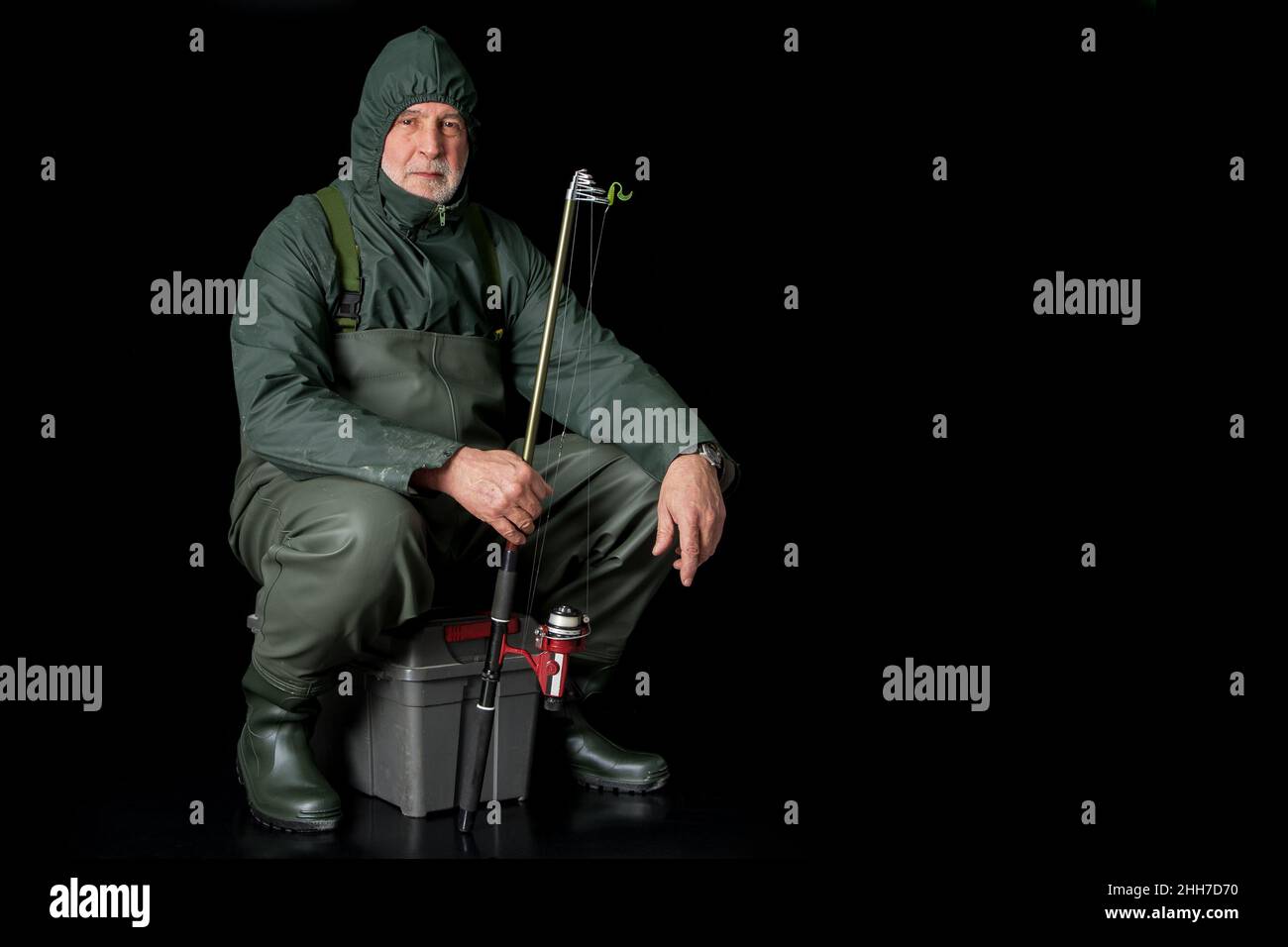 Ein Fischer in Watvögeln und Regenmantel sitzt auf seinem Angelkasten vor schwarzem Hintergrund und hält eine Angelrute in der Hand. Stockfoto