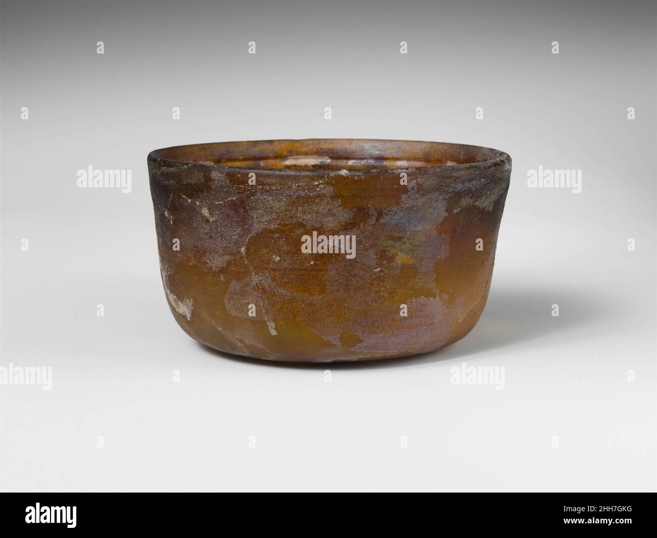 Glasschale Ende des 1st. Jahrhunderts v. Chr.–Anfang des 1st. Jahrhunderts n. Chr. römisch Durchscheinendes Goldbraun.abgeschrägter vertikaler Rand; leicht konkave Seiten, die sich nach unten verjüngen, dann zu einem fast flachen Boden gebogen.im Inneren drei horizontale Rillen mit Radschnitt: Eine breite unterhalb des Randes, die anderen zwei dünnere in einem Band um die Körpermitte; Außen schlichtes Band und flacher Grat unter dem Rand. Intakte, aber innere Risse im Boden und an der Seite; wenige Nadelstich-Blasen; stumpfend, Lochfraß und Flecken von brillanter Irideszenz und dicker cremiger Verwitterung. Glasschale 245679 Stockfoto