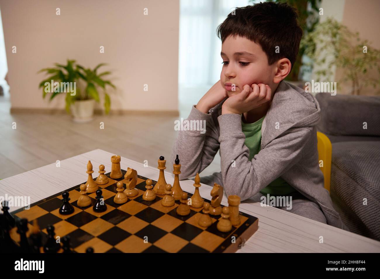 Fokussierter intelligenter europäischer Junge im Schulalter spielt zu Hause Schach, denkt und schaut sich das Schachbrett an, entwickelt eine Schachstrategie. Das Konzept der Intelligenz Stockfoto