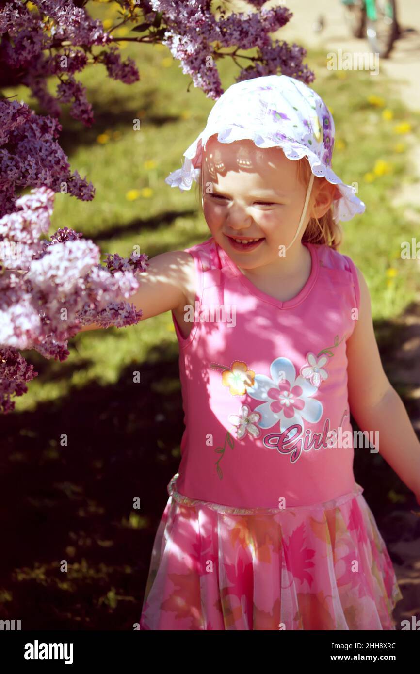 Schönes kleines blondes Mädchen mit panama auf dem Kopf, das im Frühlingsgarten mit blühendem Busch aus purpurem Flieder spielt. Stockfoto