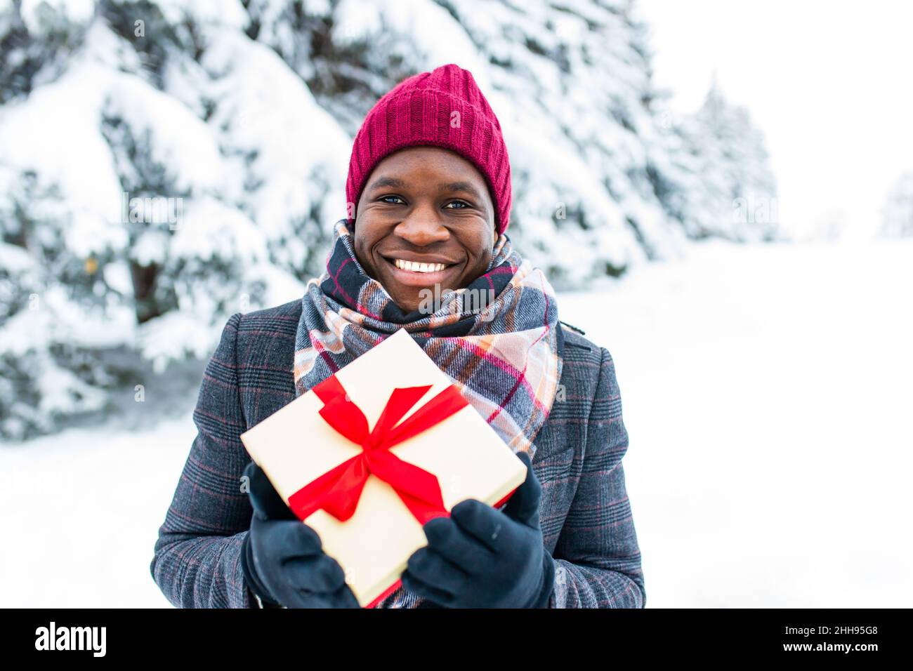 Polyethnischer glücklicher Mann in stilvollem Mantel und Hut mit Geschenkbox mit rotem Band im Winterpark, valentinstag-Konzept Stockfoto