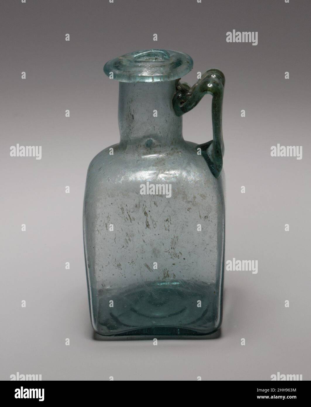 Quadratische Glasflasche 1st–3rd Jahrhundert n. Chr. römisch Durchscheinend blaugrün; Griff in der gleichen Farbe.Rand ausgeklappt, nach unten, rund und innen, bildet einen kreisförmigen, eingeengten Mund mit abgeschrägter Oberfläche; zylindrischer, aber leicht konvexer Hals mit einer horizontalen eingedrehten Einkerbung um die Basis; schräge Schulter mit abgerundeten Ecken; Quadratischer Körper mit flachen Seiten, leicht nach unten verjüngend; leicht konkaver Boden; abgeflachte zwei-Rippen-Griff als langes Pad auf der Schulter aufgetragen, vertikal nach oben gezogen, dann gebogen und nach unten, und am Hals mit nach oben Spur befestigt.auf der Unterseite Muster im Relief von Grundform, umfassend Stockfoto