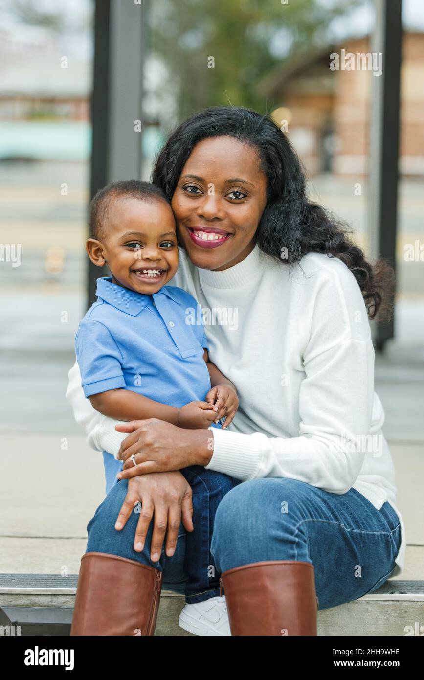 Eine wunderschöne afroamerikanische Mutter, die auf einer Treppe im Freien sitzt und ihren Sohn im Kleinkindalter umarmt und beide lächeln und sind glücklich Stockfoto