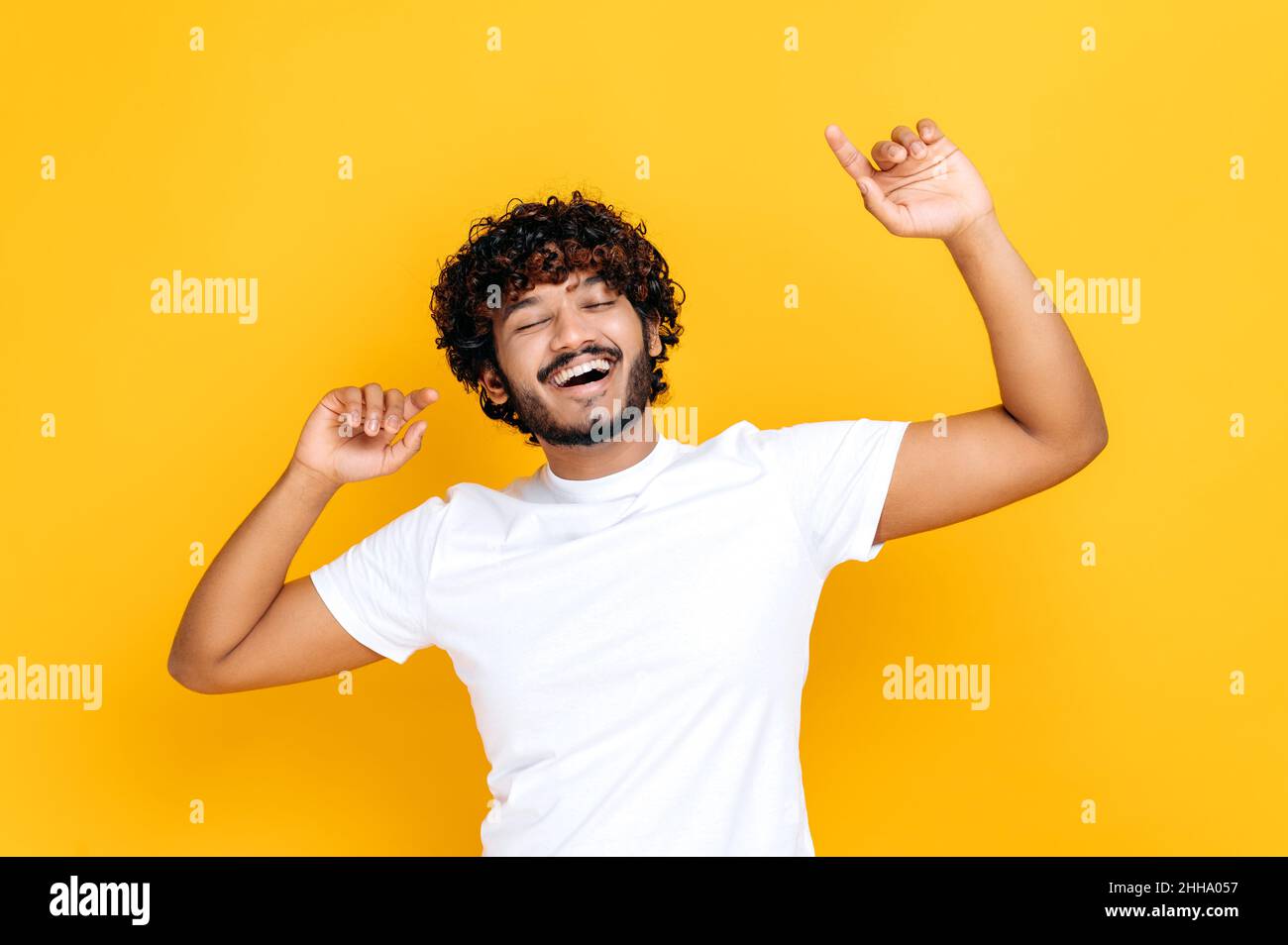 Freude, Sieg, Glück, Tanzen. Fröhlicher, fröhlicher Inder im lässigen weißen T-Shirt, tanzt zu Lieblingsmusik, hat Spaß, freut euch über Glück, feiert den Sieg, auf isoliertem orangefarbenem Hintergrund, lächelt Stockfoto