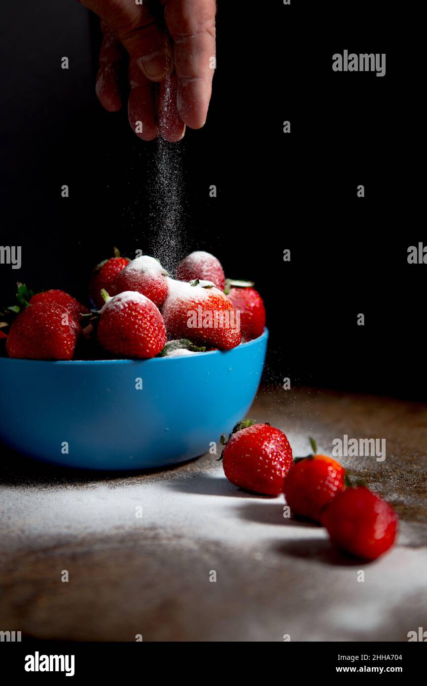 Eine blaue Schale mit frischen Erdbeeren mit einer Prise feinen Puderzuckers und dunkler und stimmungsvoller Beleuchtung Stockfoto