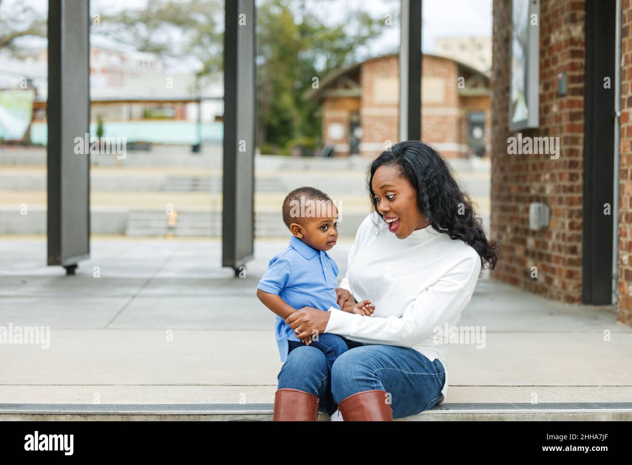 Eine wunderschöne afroamerikanische Mutter, die auf einer Treppe im Freien sitzt und ihren Sohn im Kleinkindalter umarmt und beide lächeln Stockfoto