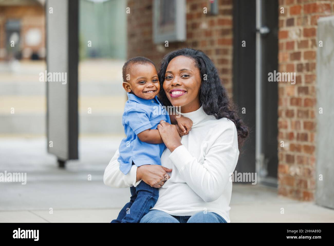 Eine wunderschöne afroamerikanische Mutter, die auf einer Treppe im Freien sitzt und ihren Sohn im Kleinkindalter umarmt und beide lächeln und sind glücklich Stockfoto