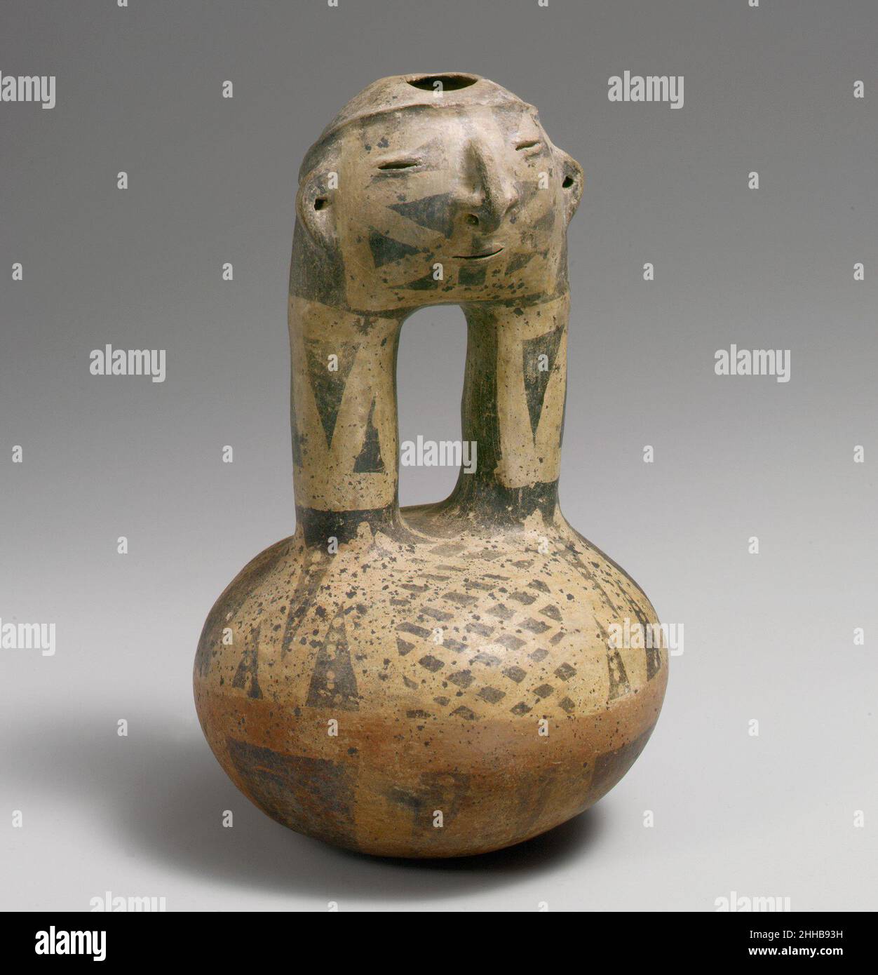 Gesichtshalsflasche 10th–16th Century Quimbaya Dieses auffällige Gefäß ist eine Steigbügelflasche mit einem Gesicht an der Oberseite der Auslauföffnungen, eine in Kolumbien relativ ungewöhnliche Form. Es wurden jedoch Beispiele unter den Grabbeigaben im Gebiet des mittleren Cauca-Flusses gefunden. Die verbundenen Ausbrüche auf diesen Flaschen bilden unweigerlich einen menschlichen Kopf mit modellierten Merkmalen, die denen der vollfigurigen Gefäße der Region ähneln. Die Flasche öffnet sich oben auf dem Kopf wie ein Beispiel in voller Figur. Die Ohren und die Nase sind durchbohrt, um Ornamente zu halten. Die dicken Ausführungen und die Kugelkammer tragen ein charakteristisches geometrisches Resist-Design Stockfoto