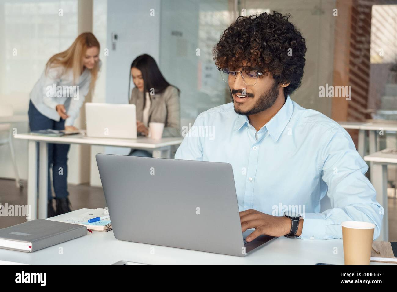 Ein vielbeschäftigter inder arbeitet am Schreibtisch am Laptop, während ein Kollege ein Brainstorming über den Hintergrund anlegt Stockfoto