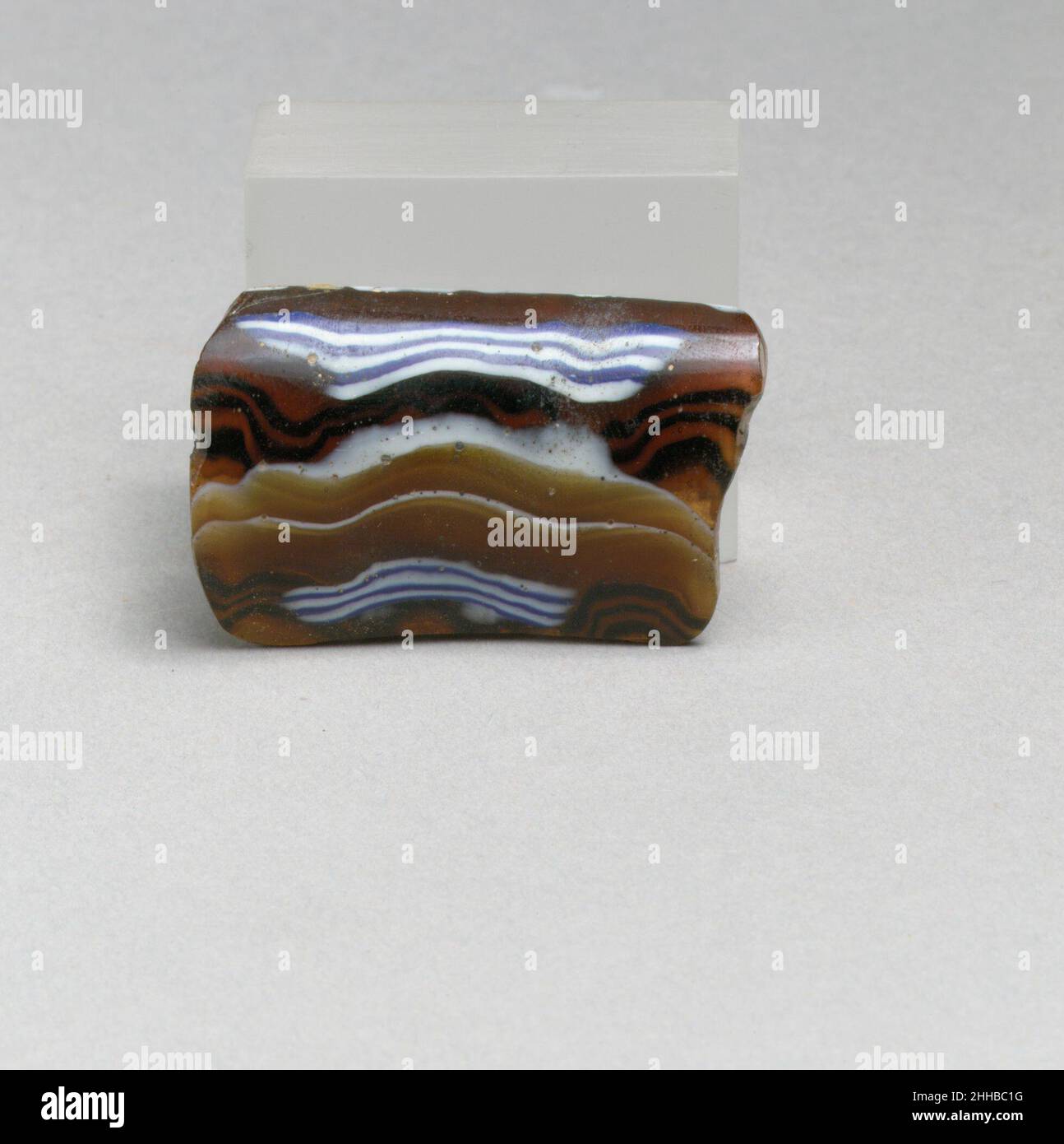 Glasmosaik geripptes Schalenfragment Ende des 1st. Jahrhunderts v. Chr.–Anfang des 1st. Jahrhunderts n. Chr. römisches Randfragment. Lichtdurchlässiges Honigbraun, Kobaltblau und opakes Weiß. Ausgespielter Rand mit dicker, abgerundeter Kante; Gerader, nach unten verjüngender Rand.Mosaikmuster aus Abschnitten eines einzelnen Rohres in braunem Boden mit unregelmäßig welligen weißen und blauen Fäden in parallelen horizontalen Linien; außen drei vertikale Rippen, weit auseinander, mit abgeflachten Oberteilen.innen poliert; innen mit Lochfraß von Oberflächenblasen; außen cremig schillernde Verwitterung; Etwas Verwitterung an zerklüfteten Rändern. Glasmosaik gerippt b Stockfoto