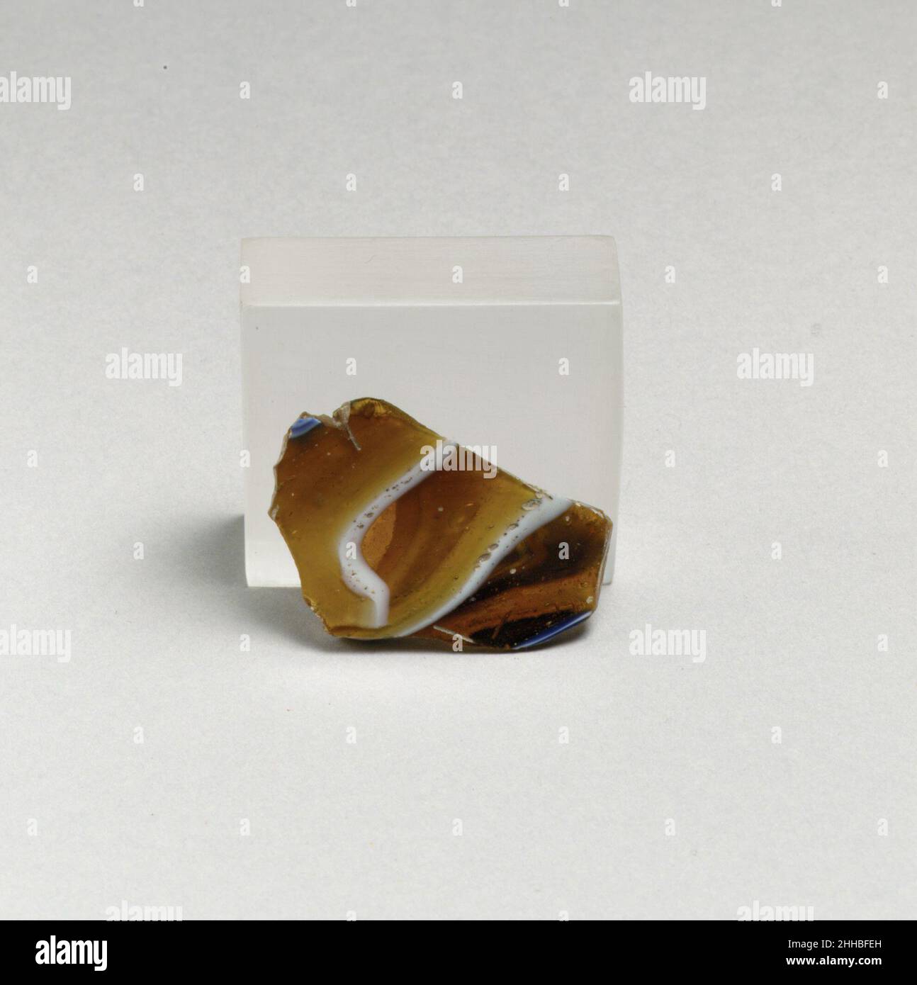 Glasmosaik geripptes Schalenfragment Ende des 1st. Jahrhunderts v. Chr.–Anfang des 1st. Jahrhunderts n. Chr. römisches Körperfragment. Lichtdurchlässiges Honigbraun, Kobaltblau, Und opakes Weiß.Fast gerade Seite, oben nach außen abfackend.Mosaikmuster aus Abschnitten eines einzelnen Rohres in einem braunen Boden mit unregelmäßigen welligen Fäden und feinen Bändern in parallelen blauen und weißen Linien gebildet; Äußerlich, einzelne vertikale Rippe mit abgerundeter Außenkante, spitz zulaufende und mit leichten Werkzeugspuren an der Oberseite.Pinprick und größere Blasen; poliertes Innere; Lochfraß von Oberflächenblasen im Inneren; stumpfende und cremig schillernde Verwitterung Stockfoto