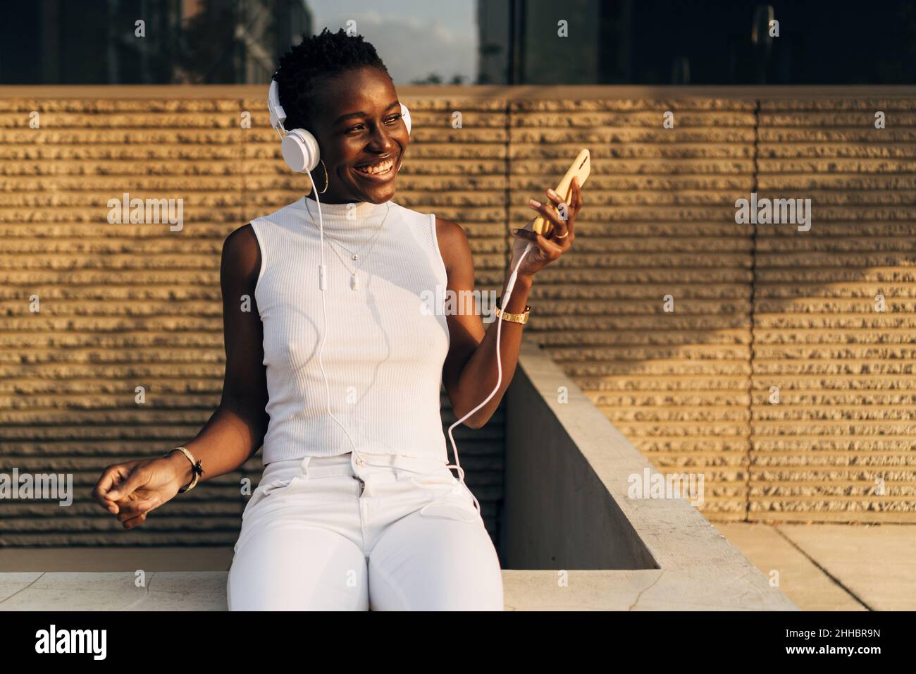 Die schwarze Frau lächelt, während sie mit einem Mobiltelefon und Kopfhörern im Freien Musik hört. Stockfoto