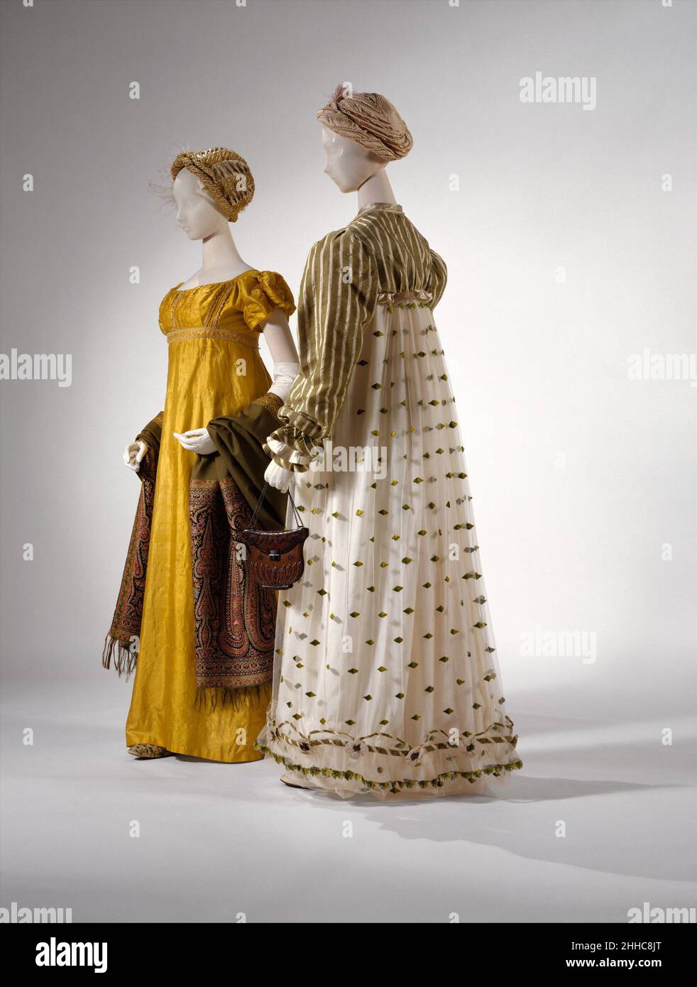 Kleid 1814 Britisch in den frühen Jahren des 19. Jahrhunderts wurde das hochtaillierte Kleid im Empire-Stil durch die Verwendung schwererer Stoffe und mehr Besatz fester. Das Kleid aus dem Jahr 1820 in der Vorderseite dieses Bildes offenbart den Impuls zur Verschönerung der einfachen Chemise der 1800s. Stickerei in Limettengrün verleiht dem Rock mehr Gewicht, während der Satin spencer eine Basis für Bänder und Rüschen bietet. Dahinter ist auch das Kleid von 1812–14 (1985.222.2a, b) fester, aus Seide gefertigt und mit Seidenspitze verziert. Die Puffärmel werden durch die Zugabe noch substantieller gemacht Stockfoto
