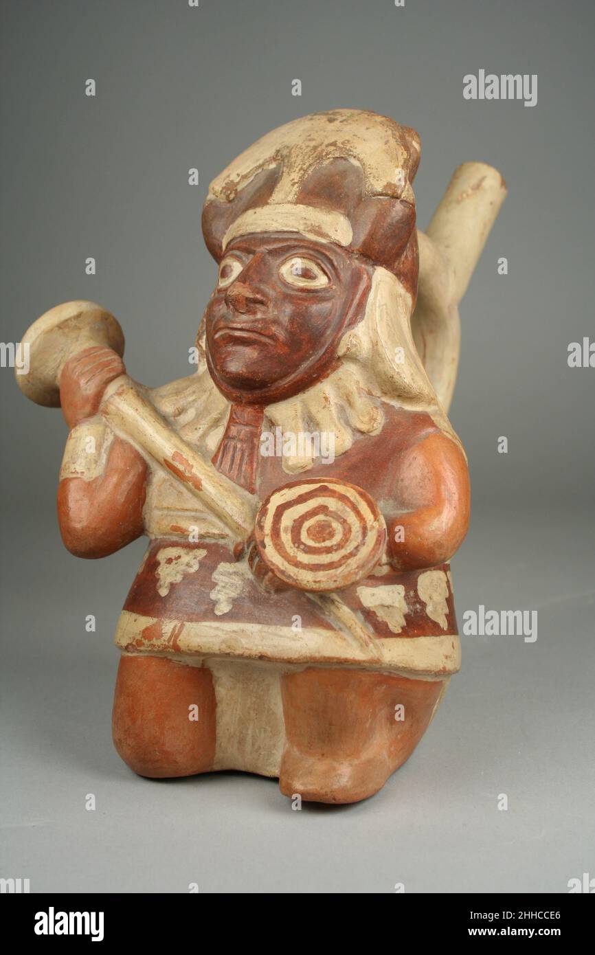 Steigbügelauslauf Flasche mit Kriegerfigur 1st–4th Jahrhundert Moche Dieses Keramikgefäß in Form eines Kriegers auf gebogenen Knien wurde von Handwerkern der Moche-Kultur der peruanischen Nordküste hergestellt. Die Figur trägt Kampfregalia, darunter einen kegelförmigen Helm, der unter dem Kinn befestigt ist und mit einer nach oben ragenden Metallsichel, einer rot-weißen Tunika, einem Gürtel, einer Halskette mit länglichen Sektkorkperlen, Handgelenkverzierungen und einem Lendenschurz. Er trägt eine Keule und einen kleinen Schild mit einem Design aus konzentrischen weißen und roten Kreisen. Das dunklere Pigment seines Gesichts deutet darauf hin, dass er Fac trägt Stockfoto