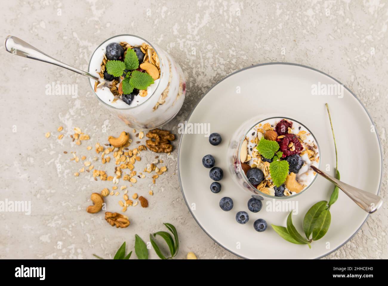 Zwei Gläser köstlicher Naturjoghurt und Müsli, mit frischen Beeren, Nüssen und Minzblättern auf grauem Betongrund. Draufsicht Stockfoto