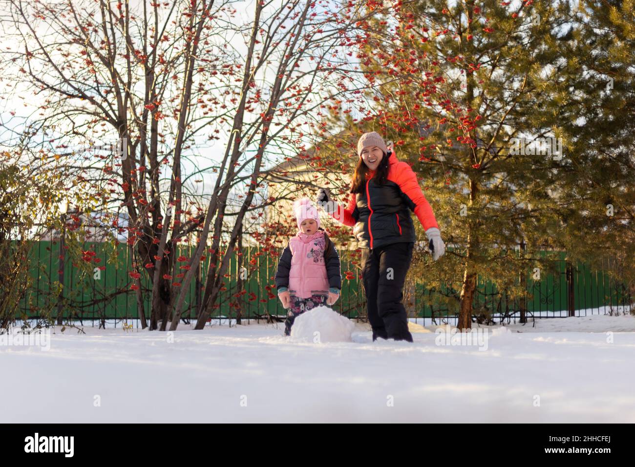 Nette kleine Tochter und Mutter Rollen große Schneebälle für den Bau Schneemann lächeln auf Hinterhof am Abend mit Eberesche und Tannen im Hintergrund Stockfoto