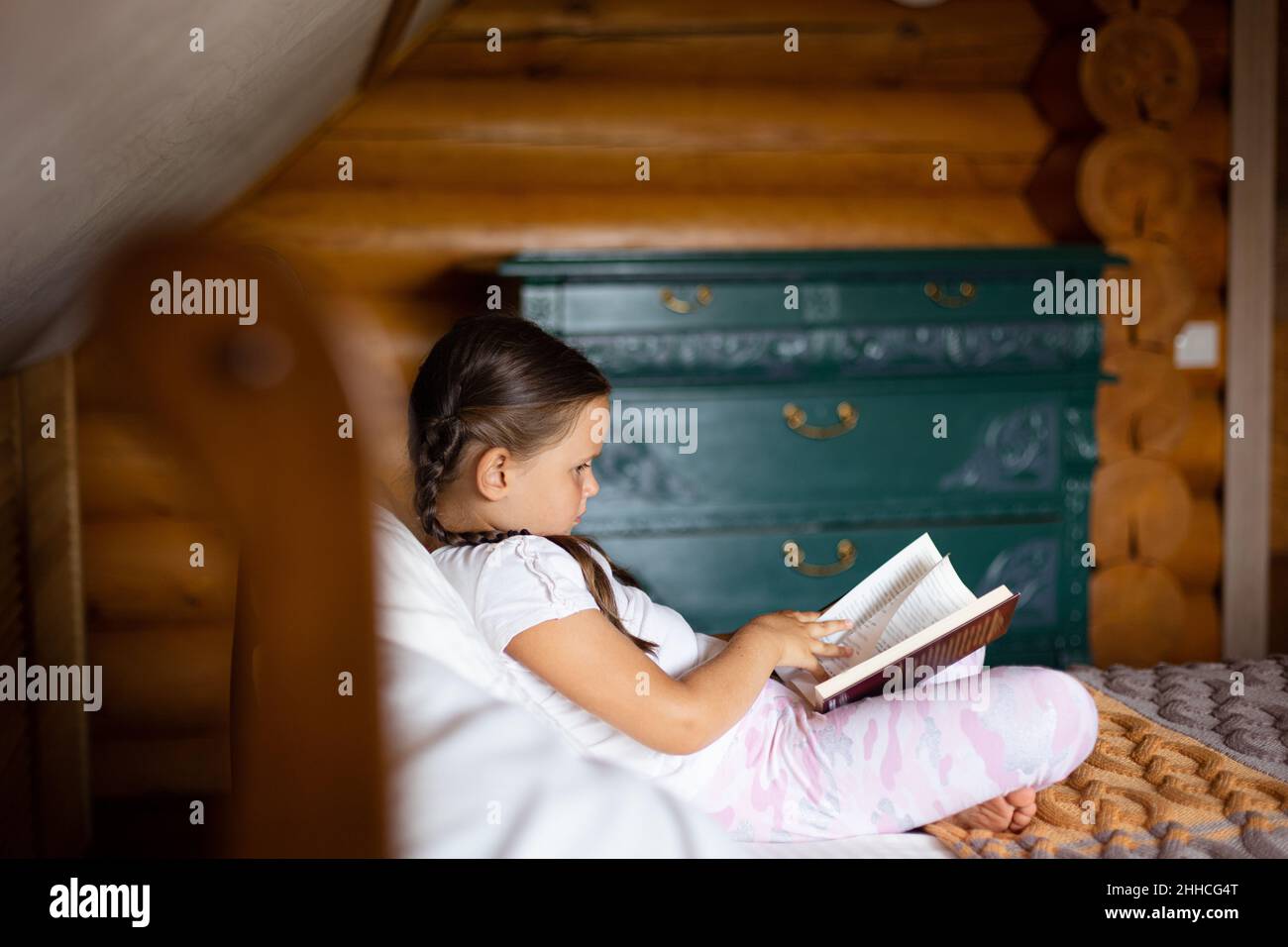 Kleines nettes Mädchen von der Seite abgebildet sitzen auf einem großen Bett mit Buch in den Händen Blick auf sie und lesen mit Kommode im Hintergrund in Holz Stockfoto