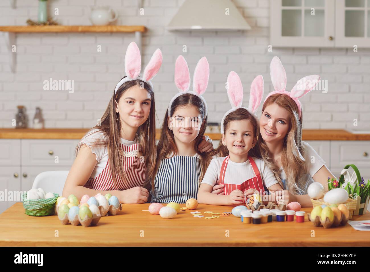 Frohe ostern. Mutter und Töchter mit Kaninchenohren schmücken Ostereier an einem Tisch in der Küche. Stockfoto