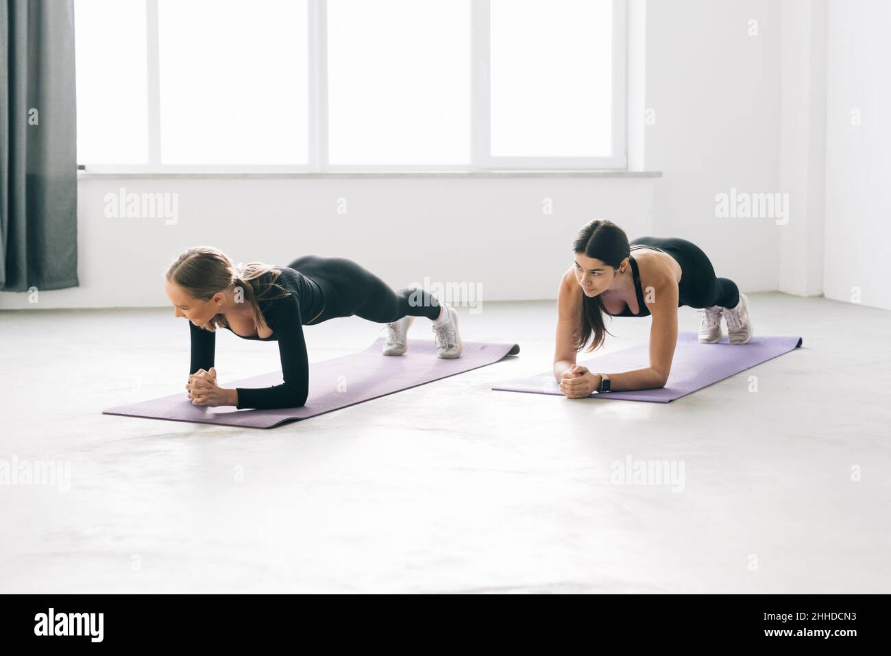 Zwei sportliche und schlanke Mädchen, die drinnen Planken machen, zusammen mit einem natürlichen Licht in der Sportstudio-Halle oder im Yoga-Kurs. Stockfoto