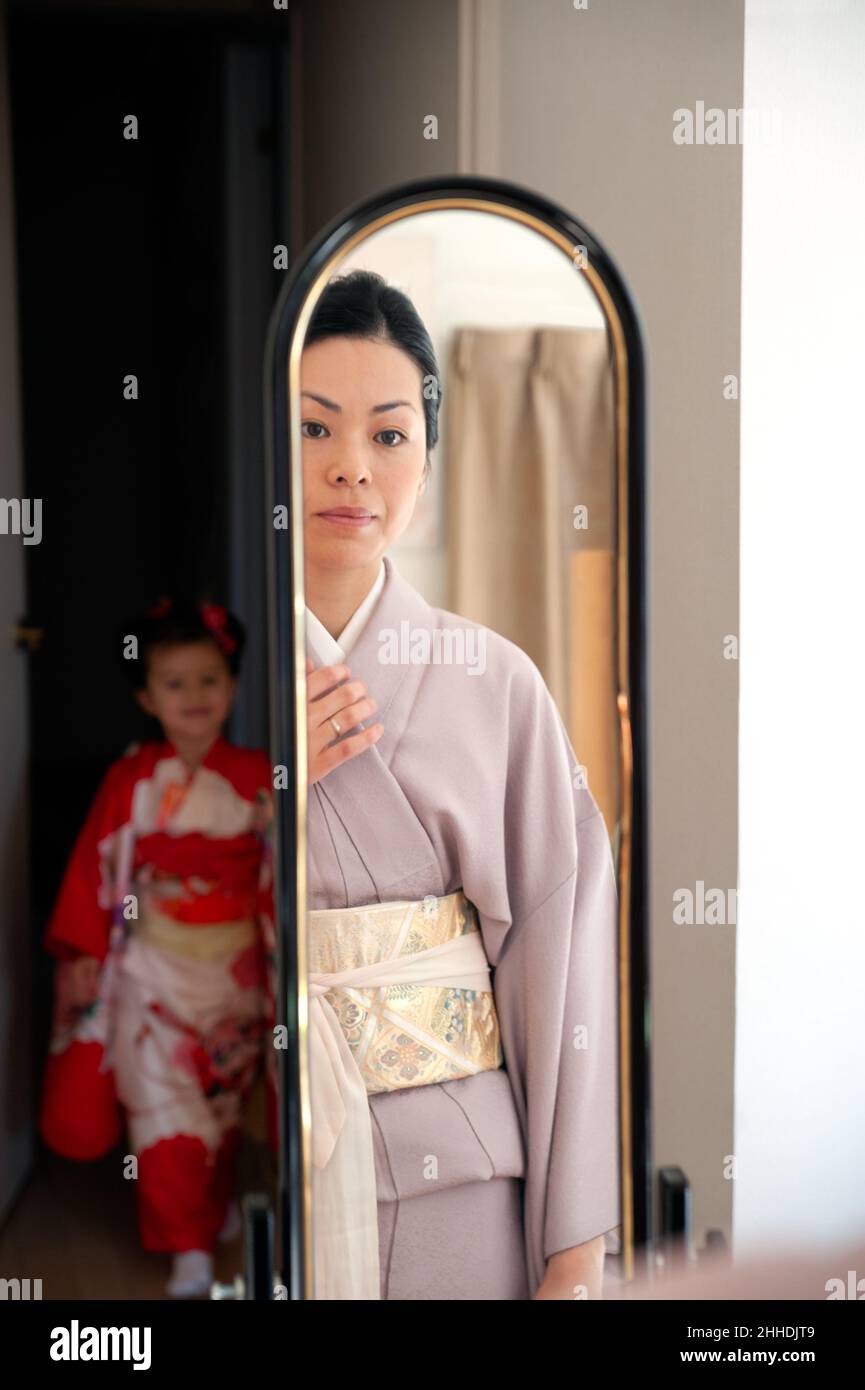 Das kleine Mädchen trägt einen roten Kimono und sieht ihrer Mutter zu, wie sie sich vor dem Spiegel in traditioneller japanischer Kleidung anzieht. Stockfoto