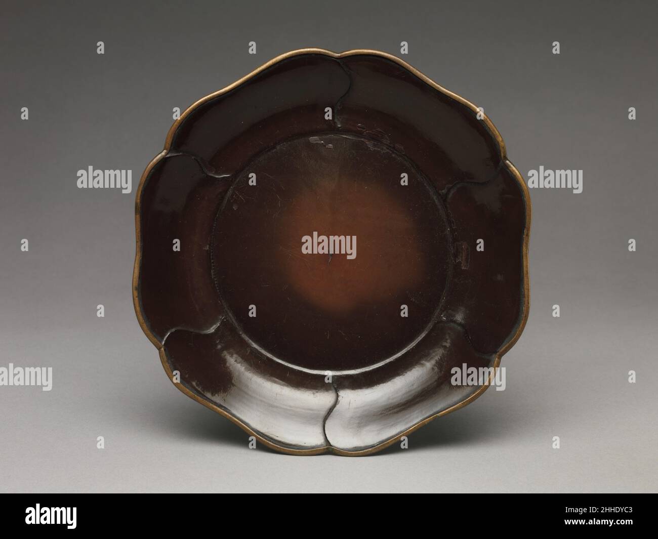 Schale mit gelapptem Rand China des 13th. Jahrhunderts in Keramik, Metallverarbeitung und Lack finden sich Schalen, die mit überlappenden Blütenblättern geformt wurden, zuerst in Nordchina um das elfte Jahrhundert, und die Form wird später zu einem Standardmotiv in der Kunst der Yuan-Dynastie (1271–1368). Dieses Gericht, das auf dem Boden eine Inschrift mit dem Namen des Herstellers oder Besitzers trägt – eine übliche Praxis während der Südlichen Song-Dynastie – sollte wahrscheinlich eine sechsblättrige Kamelie darstellen, eine Blume, die oft mit Freude und Schutz verbunden ist. Schale mit gelapptem Rand. China. 13th Jahrhundert. Lack mit Vergoldung. Südliche Song-Dynastie (112 Stockfoto