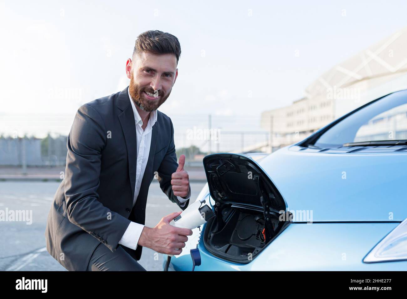 Der männliche Geschäftsmann lädt und füllt sein Elektroauto auf, schaut glücklich auf die Kamera und lächelt, zeigt den Daumen hoch Stockfoto