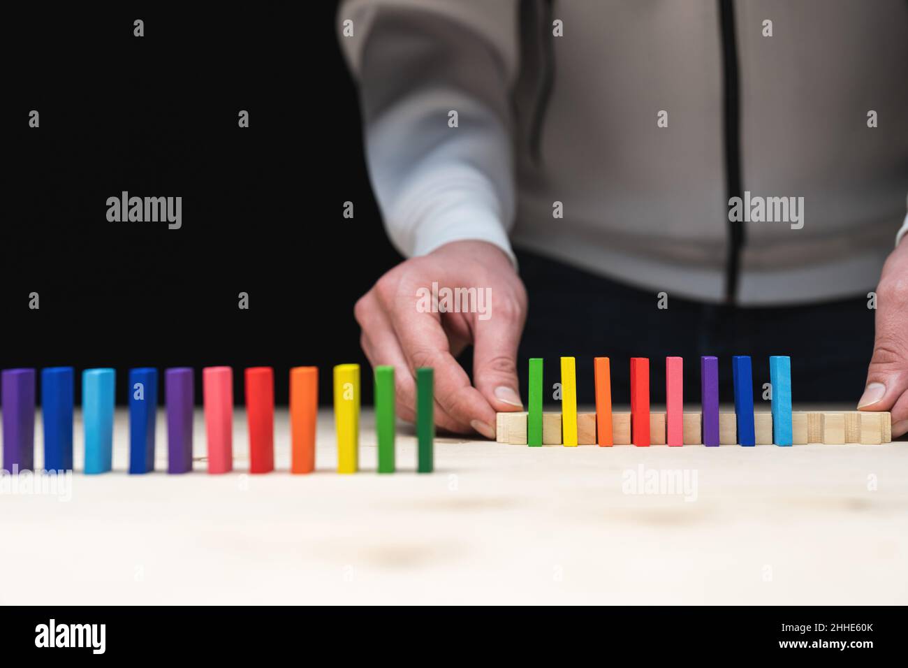 Farbige Dominosteine werden mit einem Werkzeug in der Reihenfolge der Regenbogenfarben aufgereiht. Heller Vordergrund mit schwarzem Hintergrund. Es gibt Hände im Bild l Stockfoto