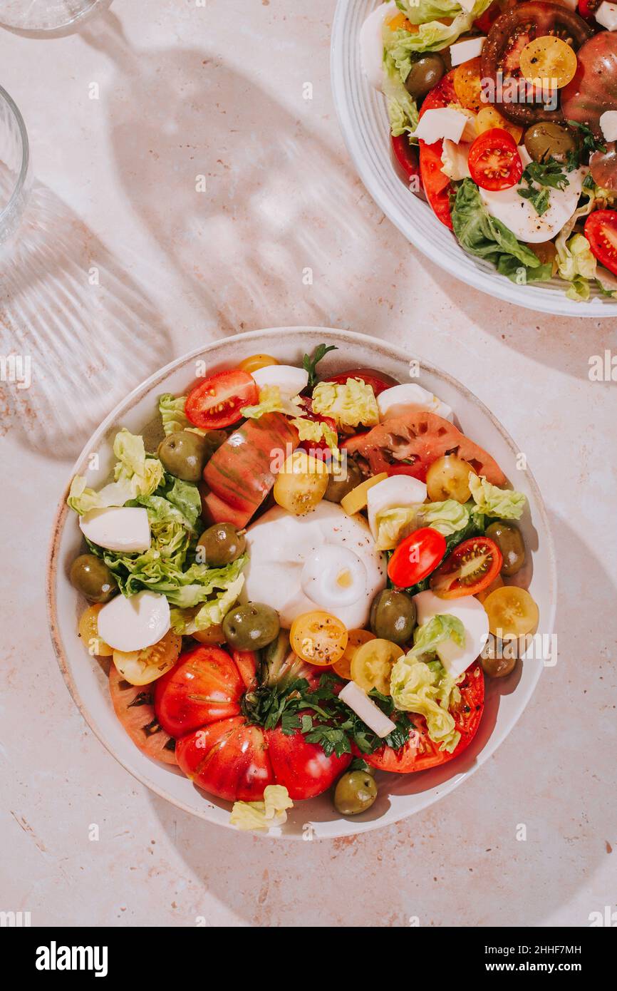 Gesunder Sommersalat mit frischen Tomaten, Burrata-Käse, Oliven und Blattgemüse auf dem Tisch mit Schatten, sonniger Stimmung, Draufsicht Stockfoto