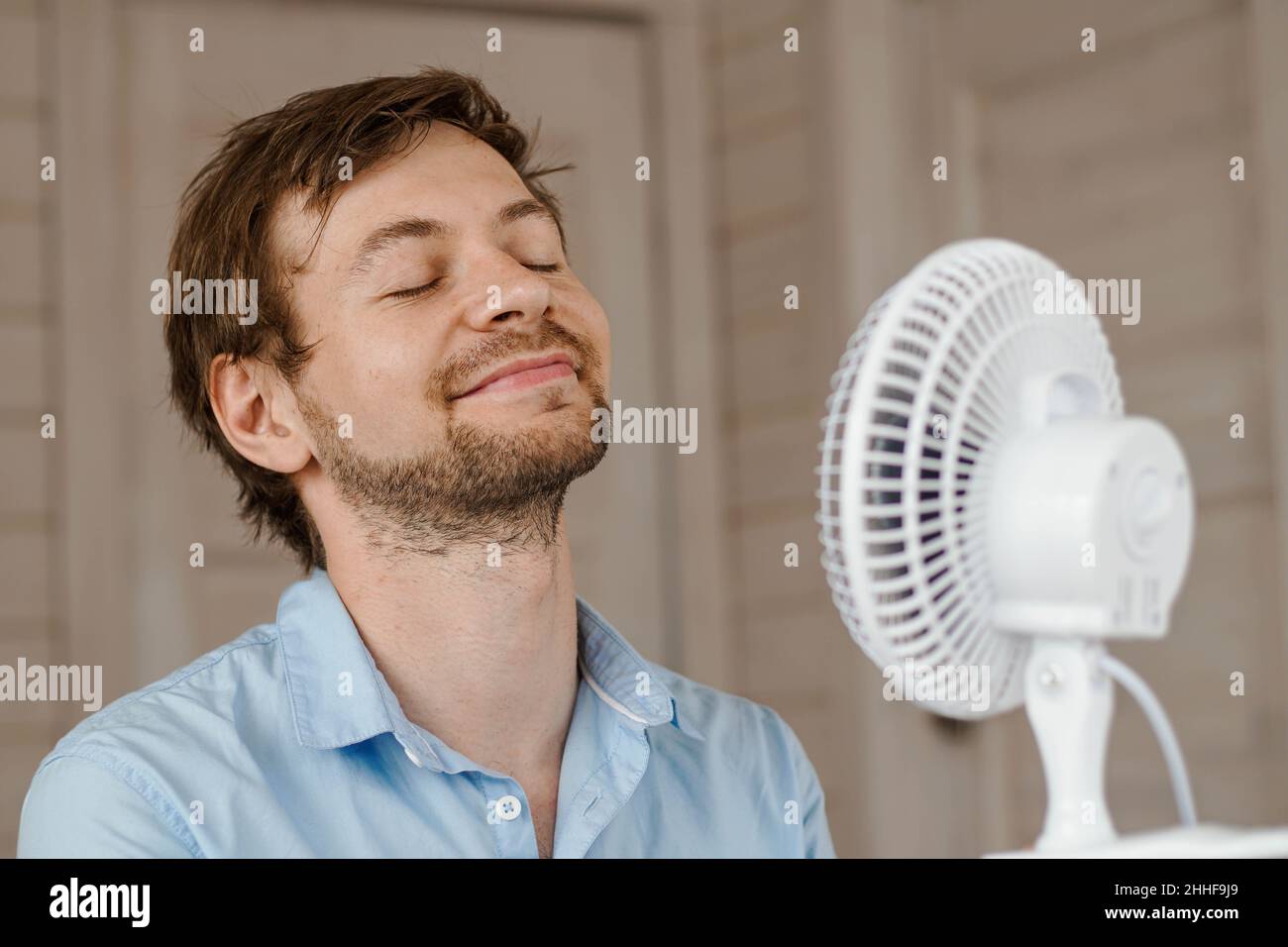 Schweißtreibender Mann, der im Büro die Luft aus dem Ventilator genießt. Geschäftsmann erfrischt sich vor einem elektrischen Luftventilator. Stockfoto