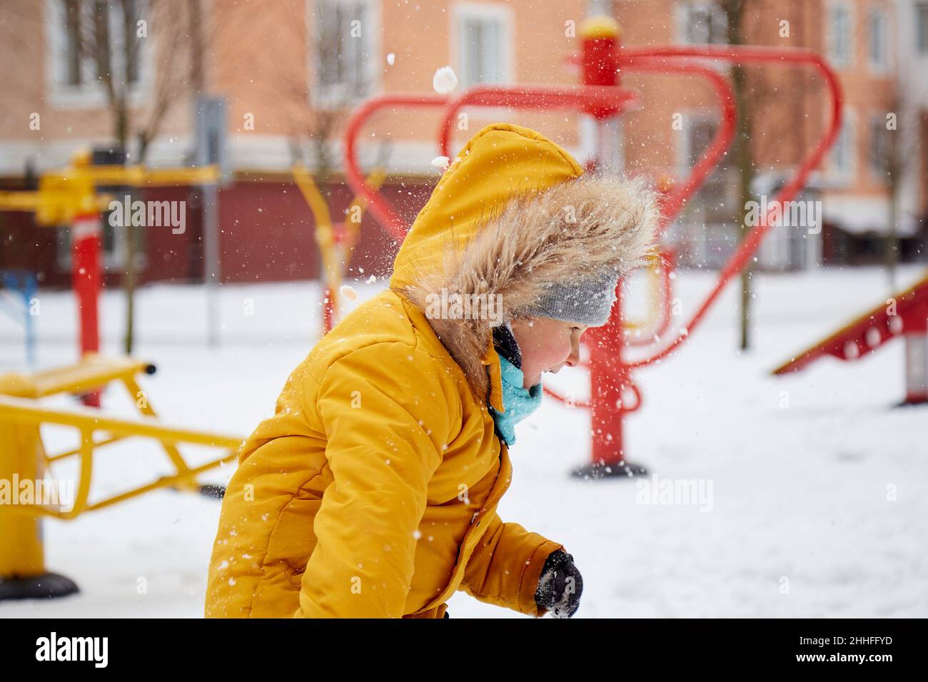 Glückliches Kind, das Spaß im Schnee im Stadthof hat. Winterspaß im Freien. Junge in leuchtend orange Winterjacke. Stockfoto