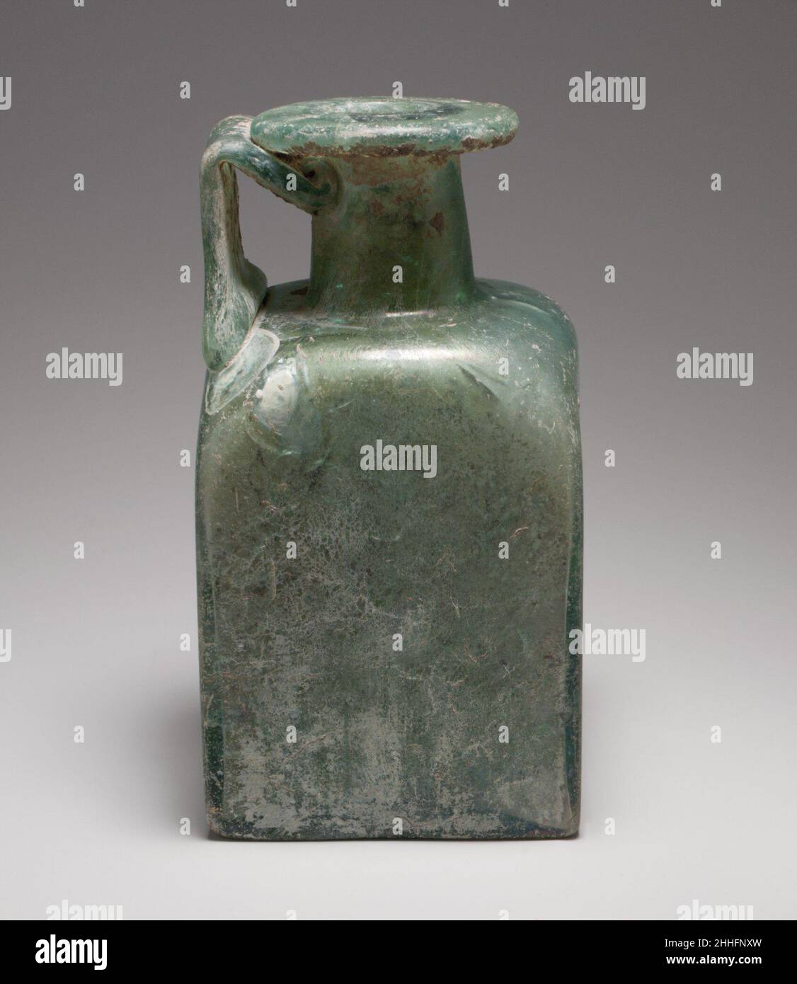 Quadratische Glasflasche 2nd. Bis 3rd. Jahrhundert n. Chr. römisch Durchscheinendes Blaugrün; Griff in gleicher Farbe; gerundeter, horizontaler Rand, über- und eingefaltet, auf der Oberseite abgeflacht, mit abgerundeter Innenlippe zu Mund; schräg, zylindrischer Hals; leicht eingeschobene Schulter, mit schräg abgerundeten Ecken; quadratischer Körper, mit flachen, vertikalen Seiten; Flacher Boden, Aber konvex auf dem Inneren; breiter, gezogener Griff, der auf einer Seite in einem langen Pad am Schulterrand befestigt ist, senkrecht nach oben gezogen, nach unten gefaltet und innen gezogen und unter dem Rand an den Hals gezogen wird.Mold-Markierung auf dem Boden, bestehend aus einem großen Kreis im Relief, der als WRE erscheint Stockfoto