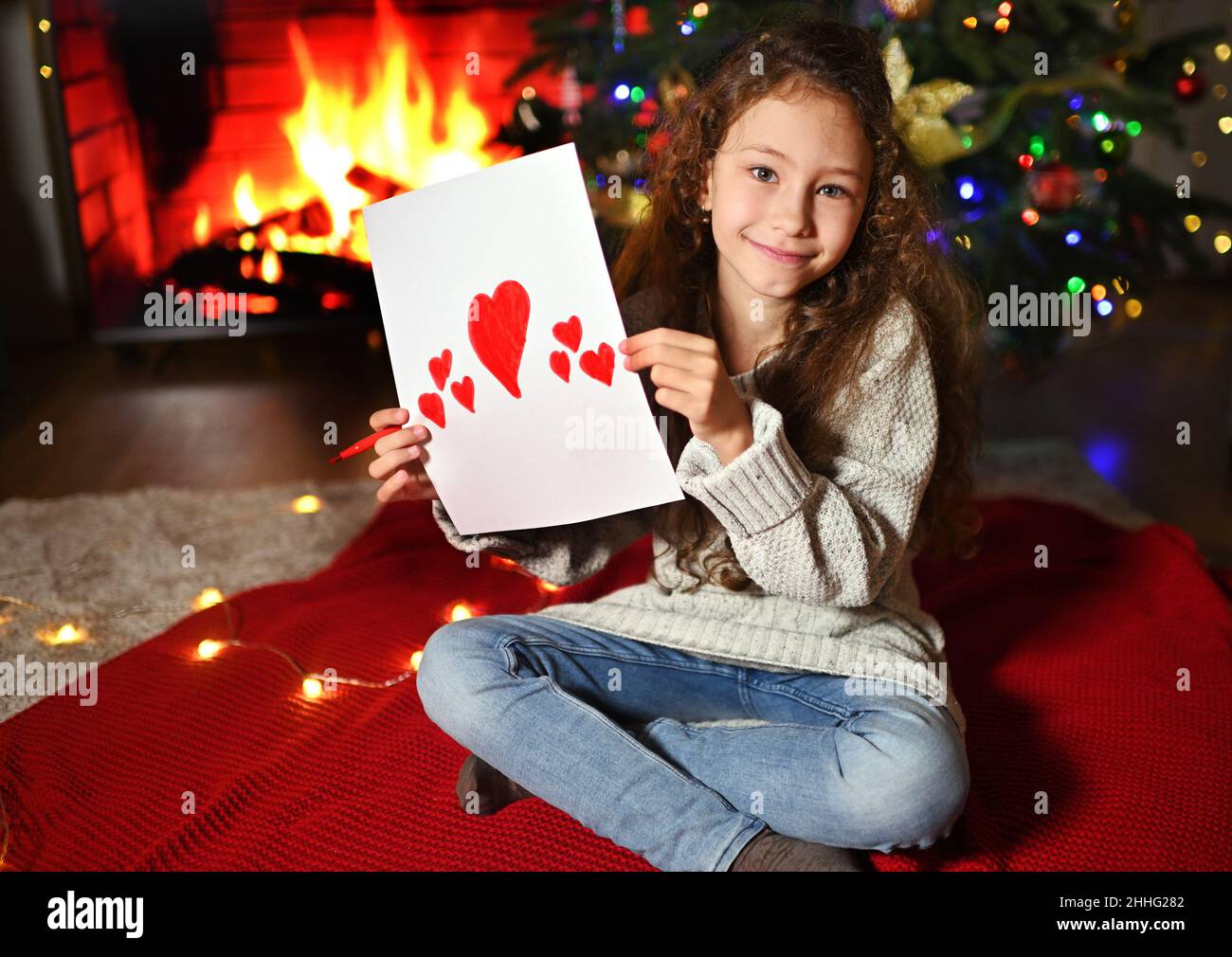 Ein nettes Mädchen mit lockigen Haaren lächelt und hält eine Valentinskarte mit roten Herzen auf dem Hintergrund des Kamins. Valentinstag Stockfoto