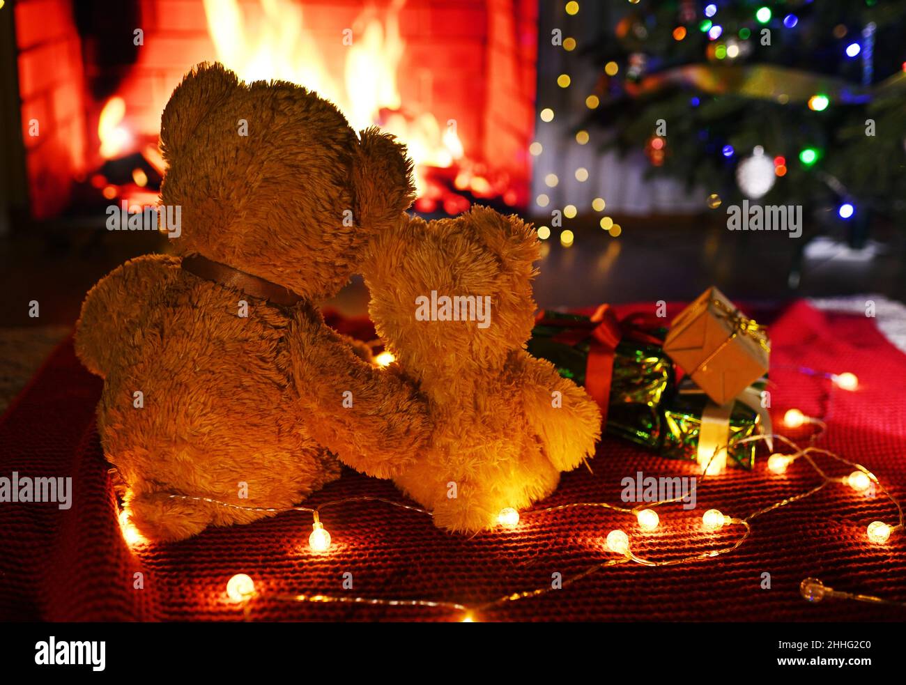 Zwei niedliche Teddybären umarmen sich gegenseitig, sitzen auf einem gestrickten roten Karree vor dem Hintergrund eines Weihnachtsbaums, von Geschenken und einem brennenden Kamin. Stockfoto