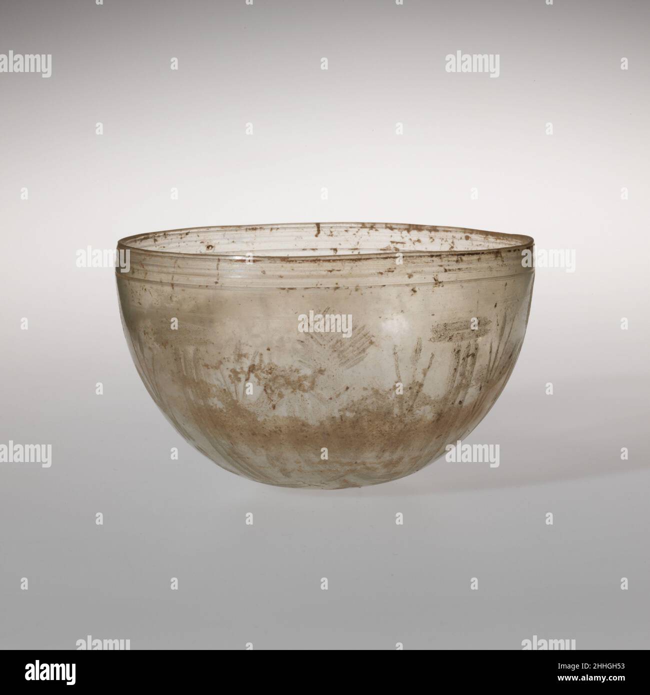 Glasschale mit geschliffenen Verzierungen 4th. Jahrhundert n. Chr. römisch farblos.unebener, leicht umgekehrter, geschliffner Rand; halbkugeliger Körper; leicht abgeflacht, aber runder Boden.Radschnitt-Verzierung außen: Unmittelbar unter dem Randband aus drei parallelen horizontalen Linien, wobei die mittlere breiter als die beiden flankierenden Linien ist; Auf dem Körper ein durchgehender Fries aus vier stehenden Figuren, abwechselnd mit vertikalen Säulen: Jede Figur zeigt nach vorne, wobei der Kopf im Profil nach links gedreht ist, eine Tunika mit vertikalen Streifen trägt und langstielige Pflanzen in beiden Händen hält, die nach beiden Seiten ausgestreckt sind, und die Säulen sind rende Stockfoto