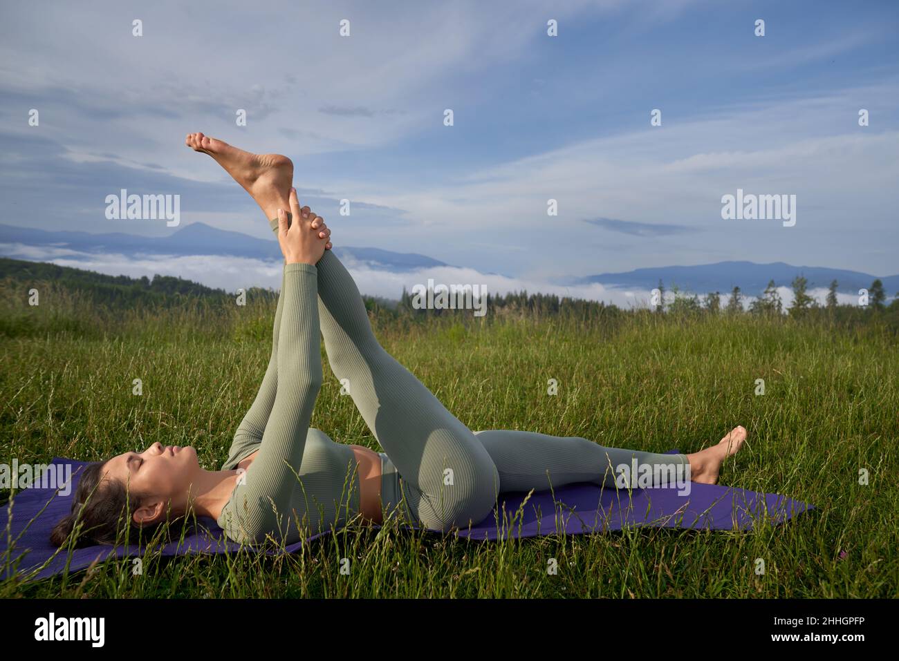 Eine athletische junge Frau macht Dehnübungen für die Beine, während sie auf einer Yogamatte inmitten grüner Berge liegt. Menschen und Outdoor-Aktivitäten Konzept. Stockfoto