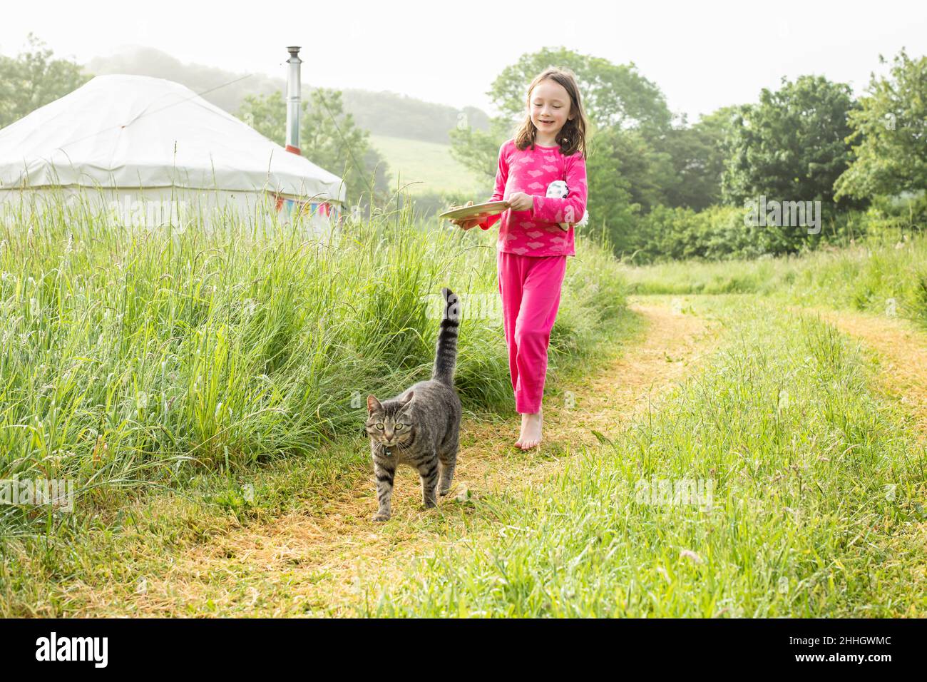 Mädchen im Pyjama zu Fuß in Grasfeld mit Katze in der Nähe Jurten auf dem Campingplatz Stockfoto