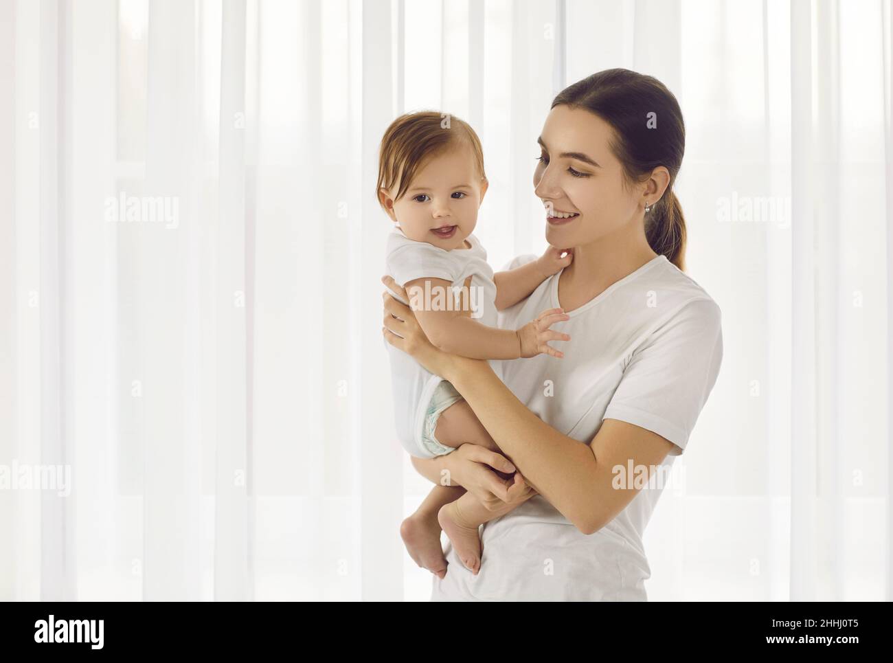 Porträt in einem hellen Zimmer mit einer jungen, liebevollen Mutter, die ihre schöne sechs Monate alte Tochter hält. Stockfoto