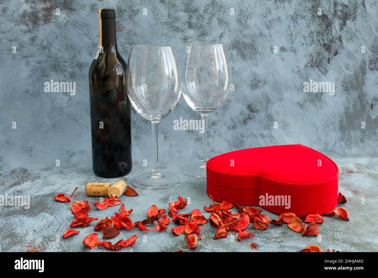 Zutaten zum Valentinstag auf blauem Hintergrund: Rotwein-Flasche, zwei Weingläser, rotes herzförmiges Geschenkkarton und trockene Rosenblätter. Seiten- oder Profilansicht. Stockfoto