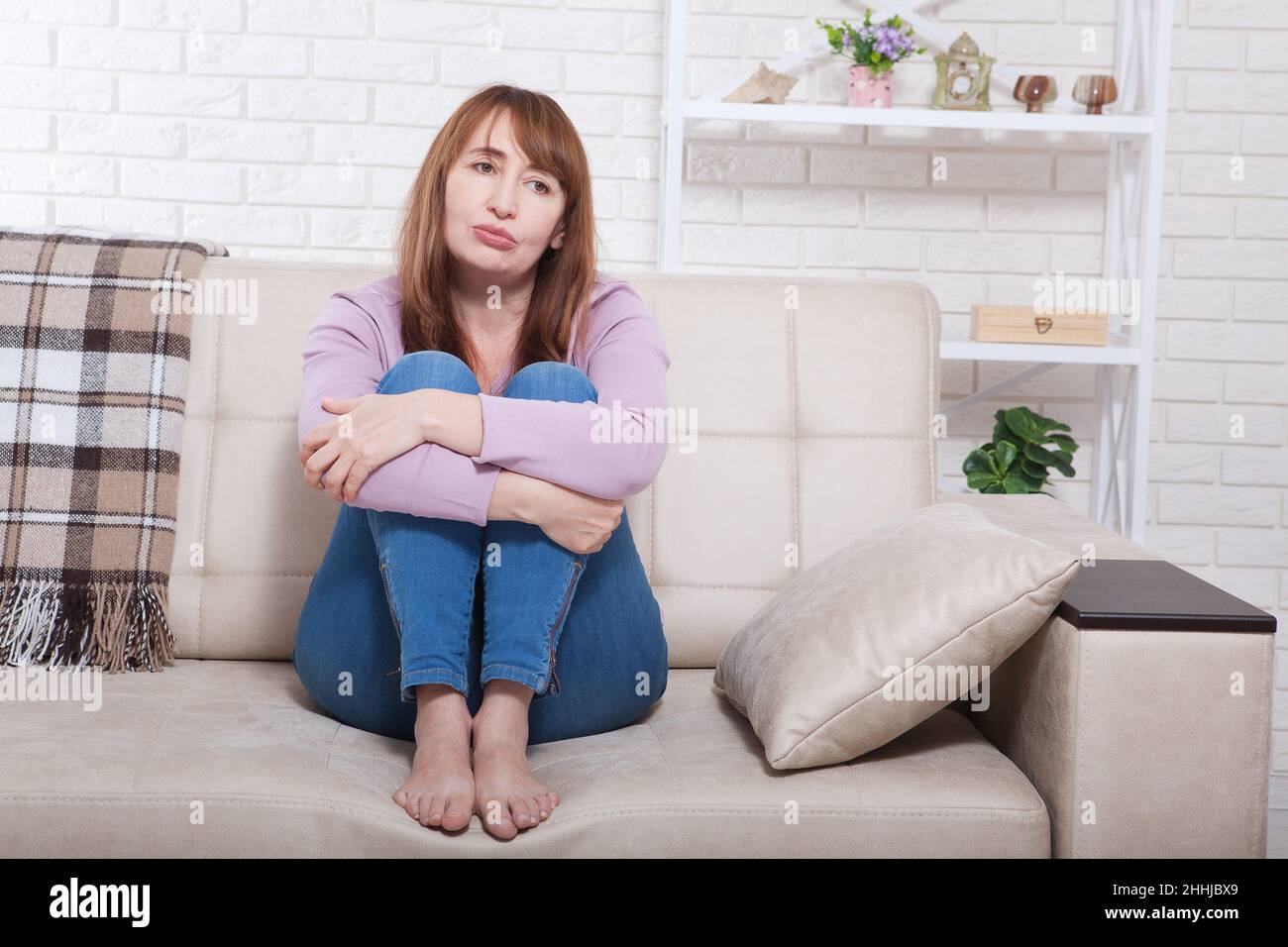 Depressive und traurige Frau mittleren Alters, die zu Hause mit geklemmten Knien auf Bett, Coach und Sofa sitzt. Speicherplatz kopieren Stockfoto