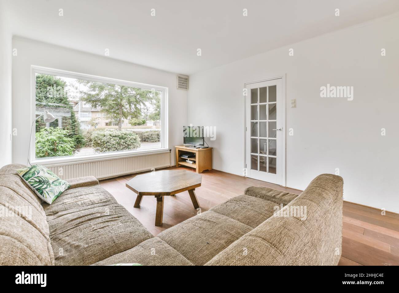 Blick auf das weiße Wohnzimmer in minimalem Stil mit Fernseher, Sofa, achteckiger Holztisch und einem großen Fenster Stockfoto