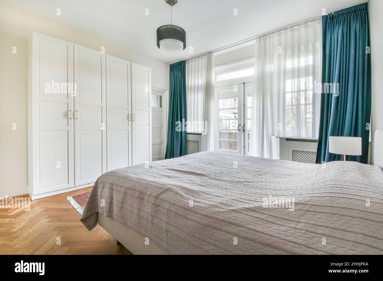 Komfortables Bett mit weißer Decke in der Nähe von Lampe und Kleiderschrank  platziert geräumiges Schlafzimmer mit geschlossener Tür und großen Fenstern  zu Hause Stockfotografie - Alamy