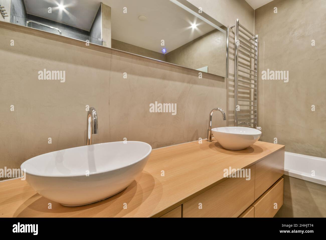Die Inneneinrichtung des Badezimmers verfügt über einen Holzschrank mit Keramikwaschbecken und Spiegel, der an der beigefliesten Wand hängt, sowie über der Badewanne beheizte Handtuchhalter aus Chrom Stockfoto