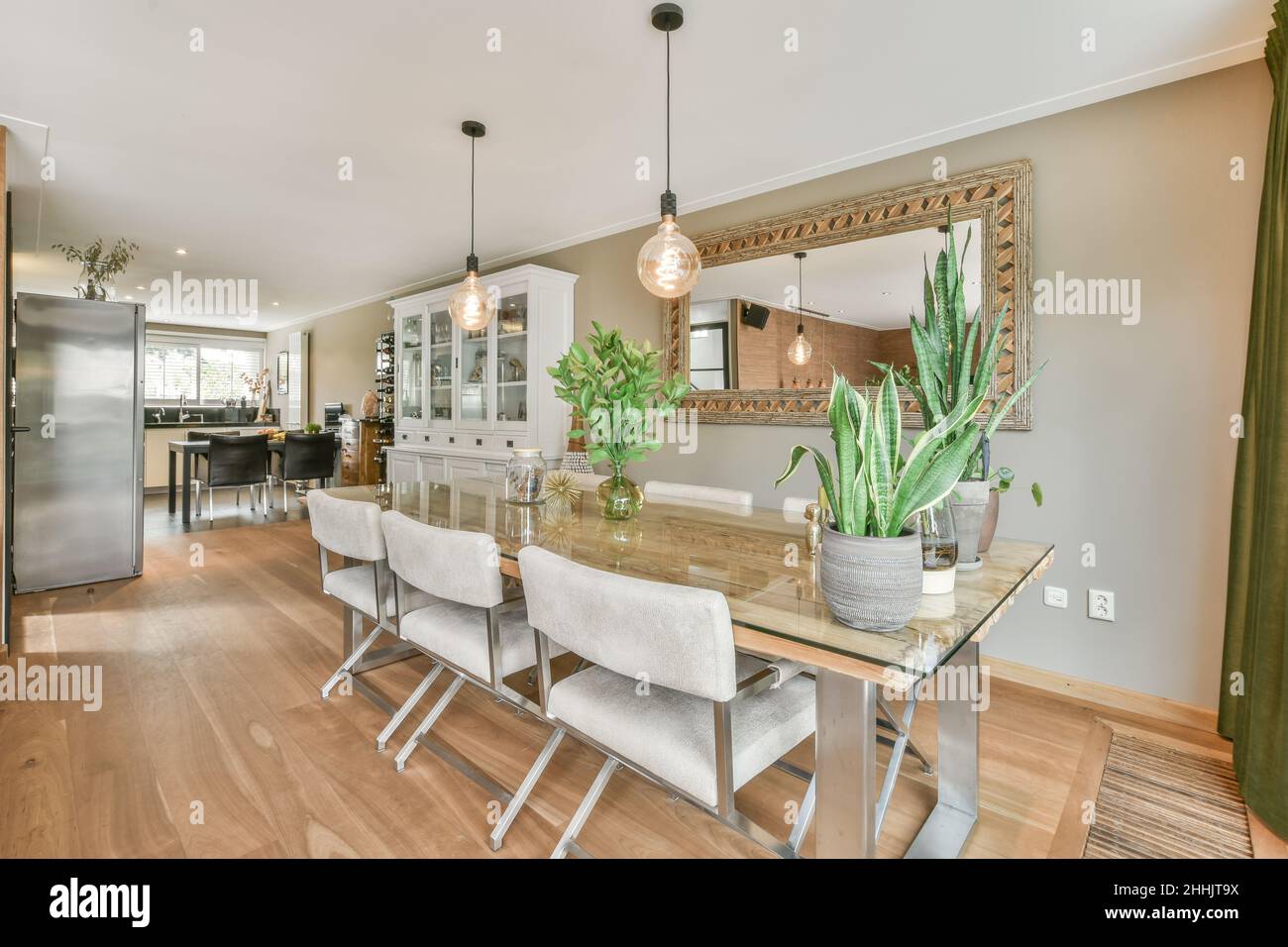 Großer Esstisch mit beigefarbenen Stühlen in einem geräumigen Zimmer, das mit einem Spiegel an der Wand und kreativen Lampen in der Nähe der Küche in einer geräumigen Villa ausgestattet ist Stockfoto