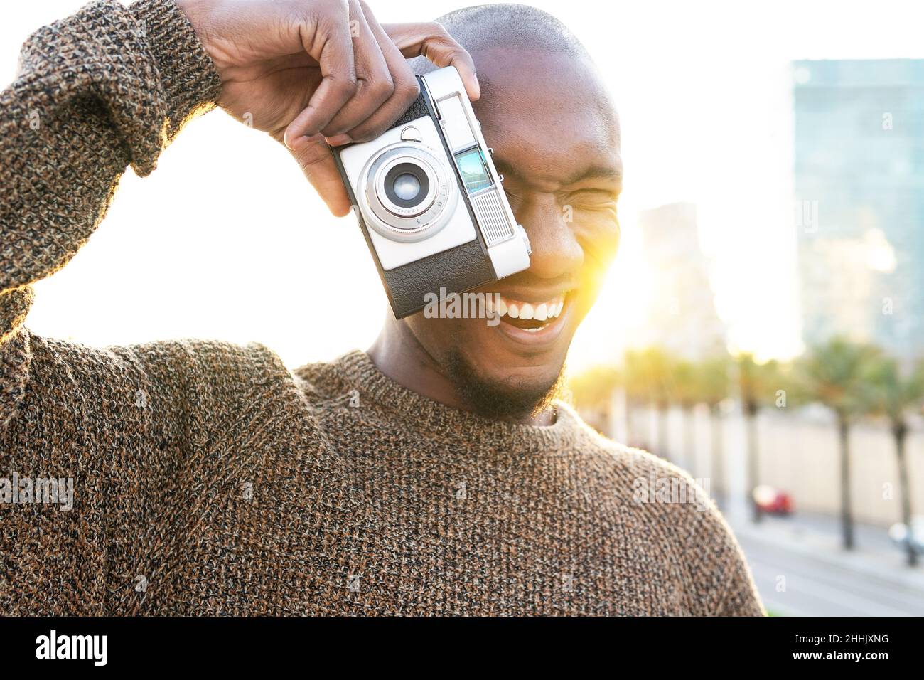Fokussierter, fröhlicher, kahlköpfiger afroamerikanischer Fotograf, der mit einer Fotokamera fotografiert, während er auf einem Rasen in der Stadt mit Bäumen steht Stockfoto