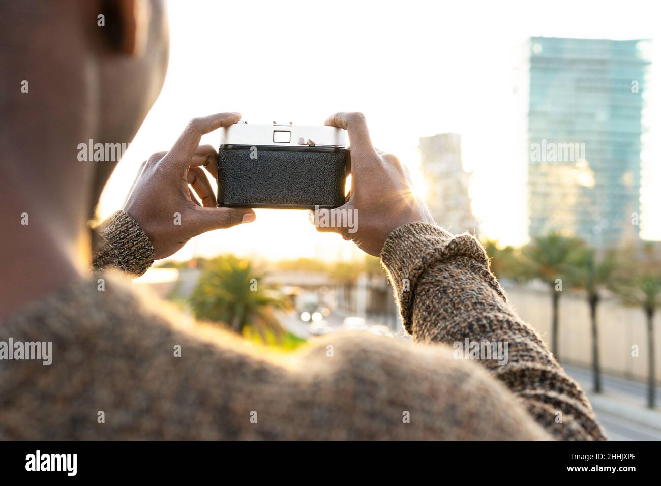 Rückansicht eines nicht erkennbaren kahlen afroamerikanischen Fotografen, der mit der Fotokamera fotografiert, während er auf einem Rasen in der Stadt mit Bäumen steht Stockfoto