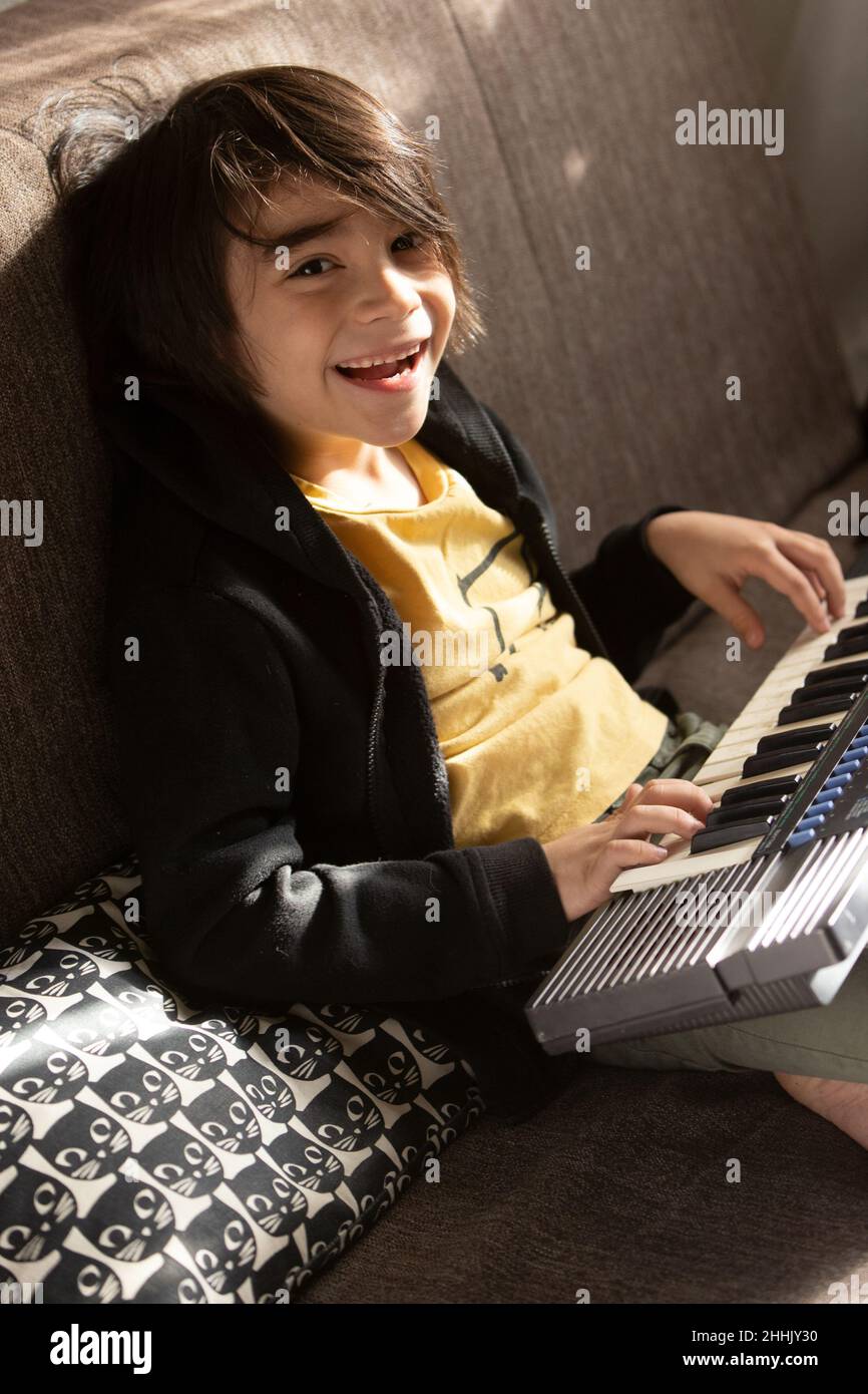 Positiver ethnischer Junge, der mit einem Lächeln auf die Kamera blickt, während er Melodie auf einem Synthesizer auf einer bequemen Couch im hellen Wohnzimmer spielt Stockfoto