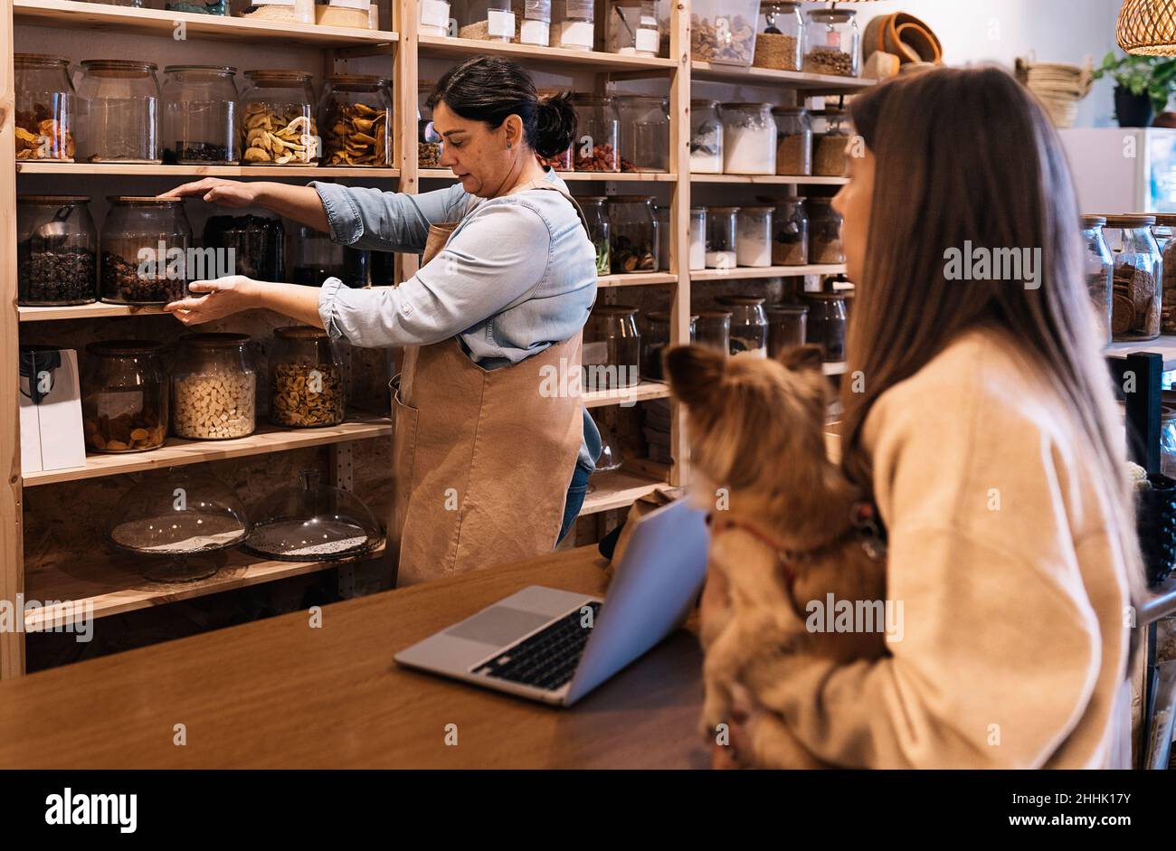 Seitenansicht einer ruhigen Verkäuferin im Vorfeld und Demonstration eines Glasglases mit Nüssen gegenüber einer Kundin, die mit Hund in der Hand an der Holztheke in der Hand stand Stockfoto