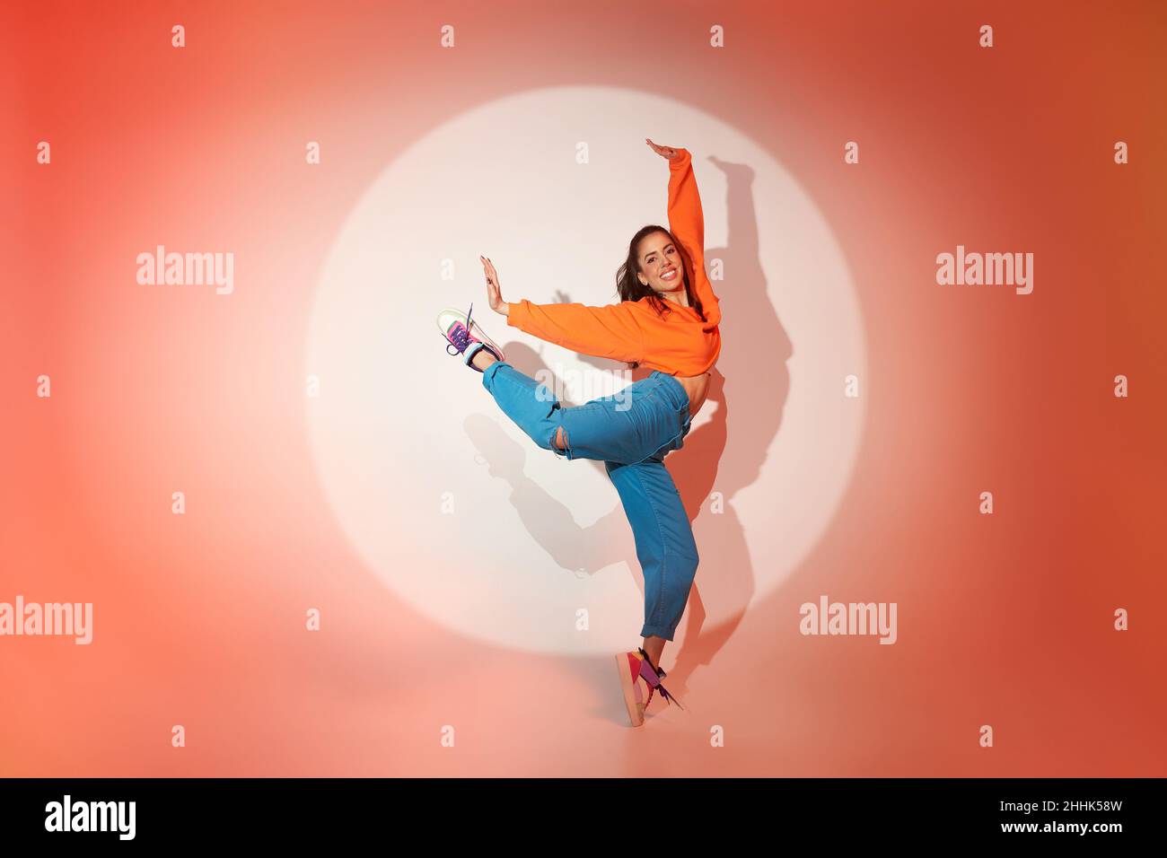 Energisch glückliche Tänzerin, die mit erhobenem Bein und ausgestreckten Armen im Licht des Projektors im Studio springt und die Kamera anschaut Stockfoto
