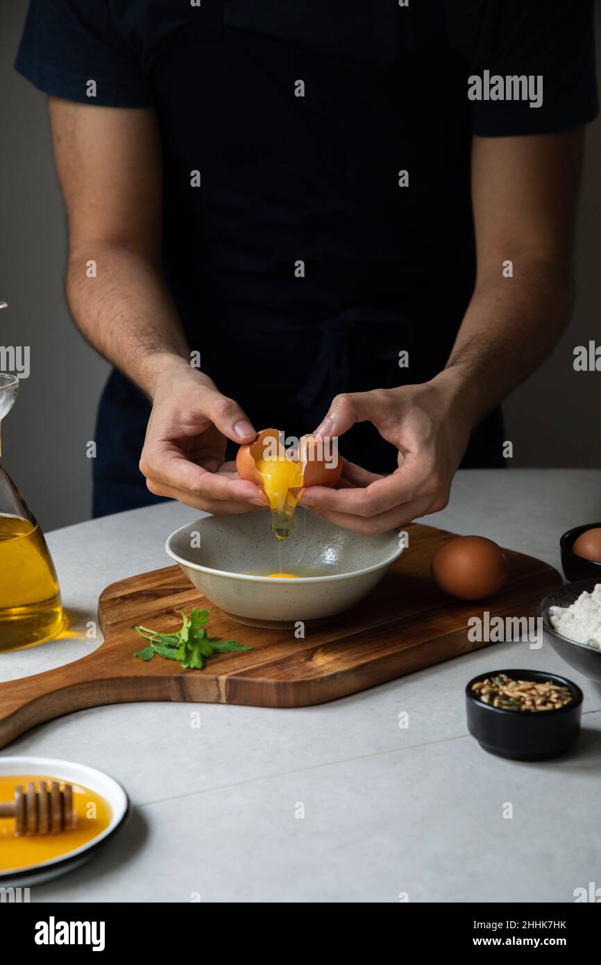 Crop gesichtslosen männlichen Koch zerbrechen rohes Ei in Schüssel auf Schneidebrett beim Kochen am Tisch mit verschiedenen Zutaten in der Küche Stockfoto